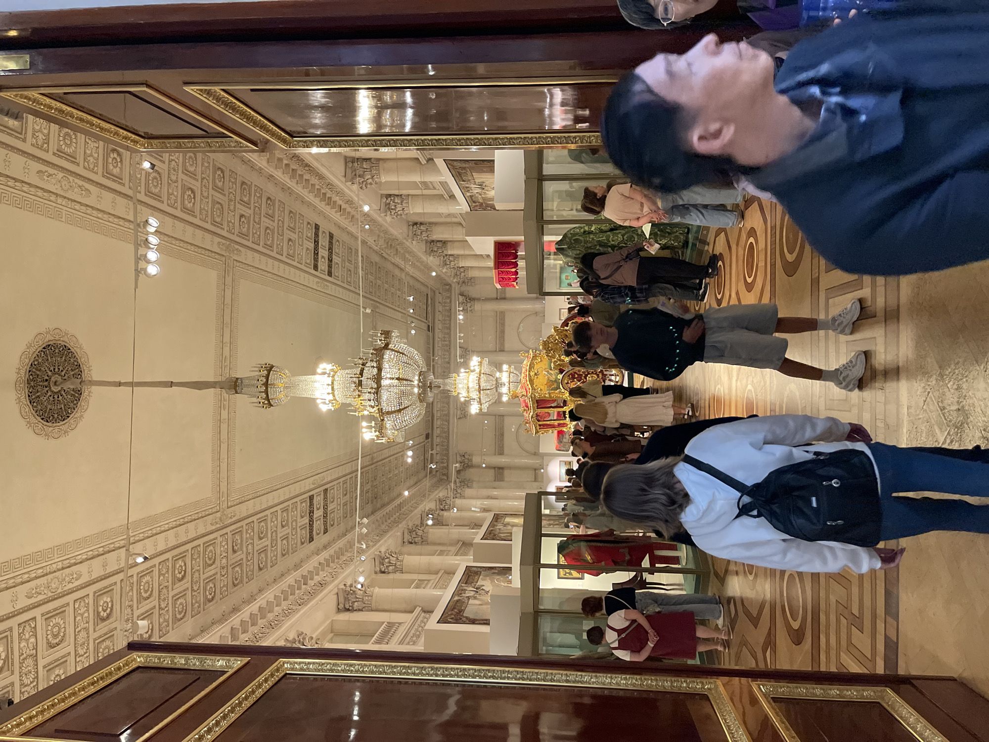 The image depicts the interior of a grand hall, likely a museum or palace, located in Saint Petersburg, Russia. The hall is characterized by its opulent architecture, featuring ornate ceilings with intricate moldings and recessed panels, as well as large, crystal chandeliers that cast a warm light. Fluted columns line the walls, supporting the high ceiling. Several people are visible, mostly seen from behind, walking through the hall. They appear to be tourists or visitors.
In the mid-ground, a crowd is gathered, with some individuals looking at exhibits. One prominent object in the distance is a decorative, gilded carriage, likely an exhibit. Display cases containing artifacts are also visible. The floor is patterned with a light-colored inlay on a darker wood, creating a geometric design. The overall atmosphere suggests a public space of historical and cultural significance, commonly found in Saint Petersburg's numerous palaces and museums. The scene is well-lit, with the chandeliers and ambient lighting illuminating the space and its occupants.
