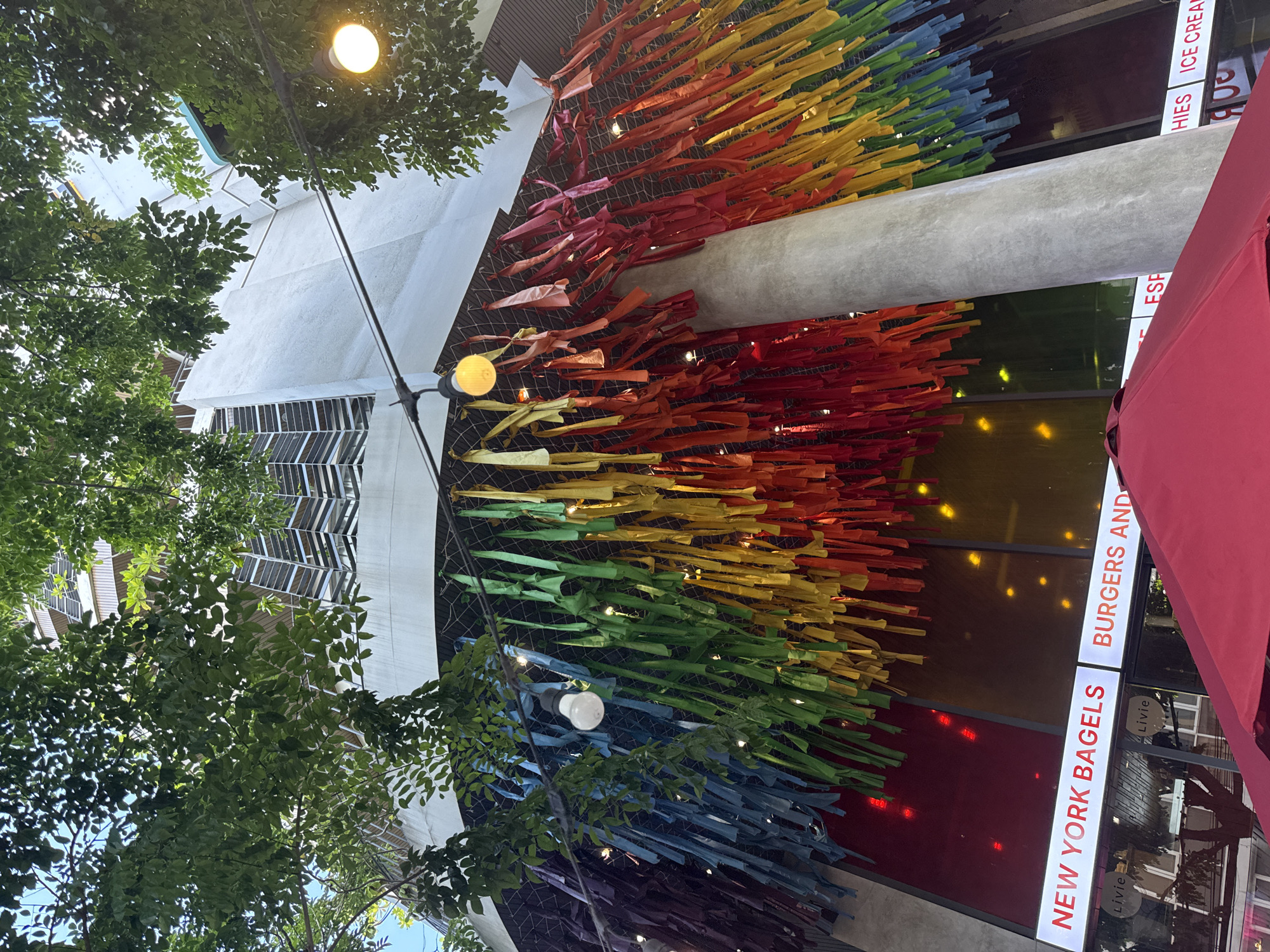 The image shows the exterior of a New York Bagels restaurant in Đà Nẵng, Vietnam. The restaurant's facade features a colorful art installation resembling a rainbow, made of strips of fabric or similar material attached to a mesh grid. The colors progress from red at the top, transitioning through orange, yellow, green, blue, and purple towards the bottom.
The restaurant's signage, partially visible on the right side of the image, reads "NEW YORK BAGELS" and "BURGERS AND." Other text, cut off in the image, includes "ESP," "HIES," and "ICE CREAM."
The surrounding environment includes trees, street lamps, and architectural elements of the building and nearby structures. A red umbrella is partially visible on the right side.