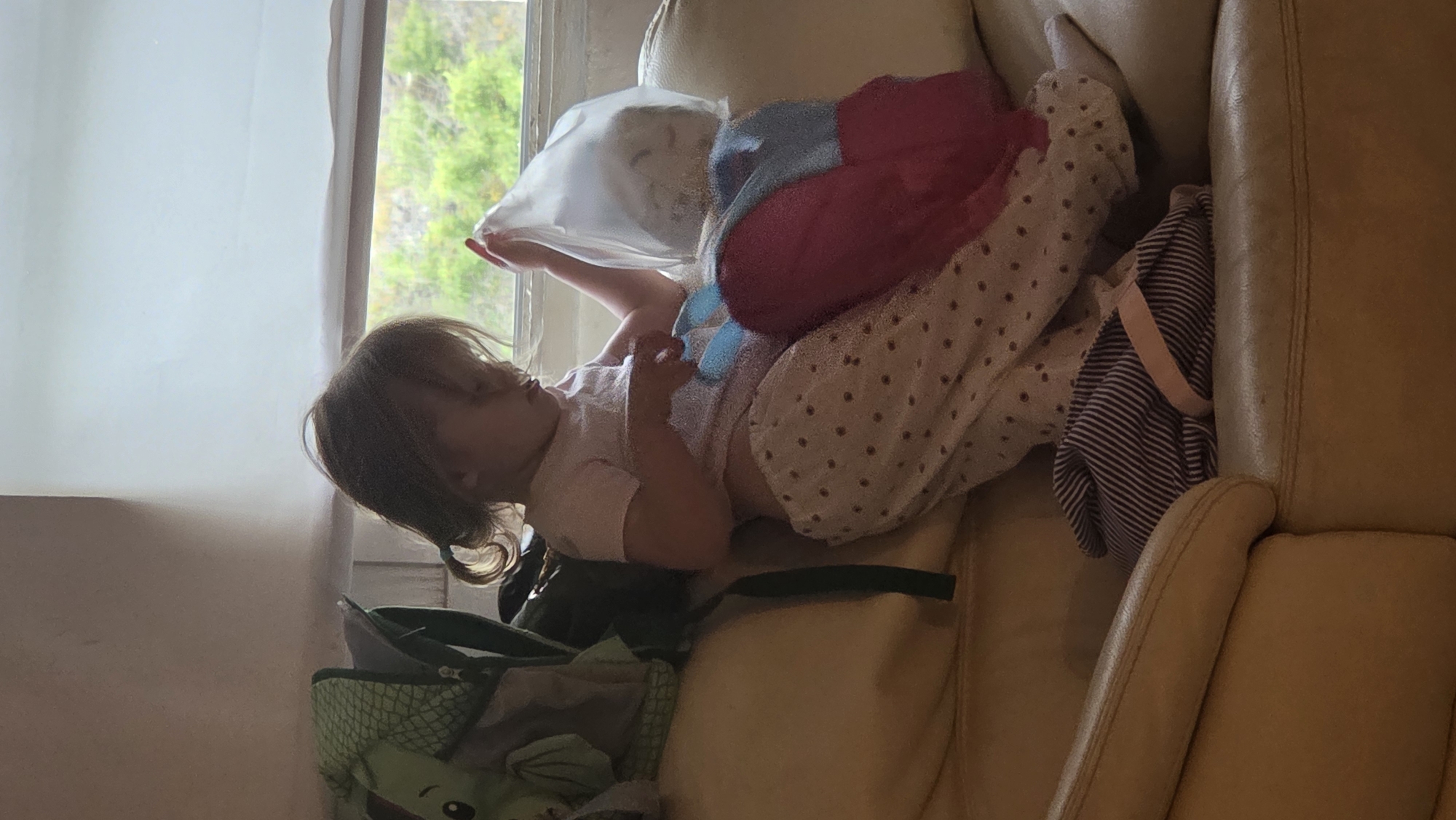 A young child is sitting on a tan sofa next to a window. The child is wearing a light pink shirt and patterned pants, and has their hair pulled back in two pigtails. They are holding a clear plastic bag in their right hand and are looking down at it. A pink and blue blanket is draped over their lap, and a striped bag is on the sofa to their left. A green backpack with a dinosaur design is on the sofa behind the child. The window shows a blurry view of green foliage and a cloudy sky. This scene is likely taking place in a residential setting in Guía de Isora, Spain, given the natural light suggesting daytime and the window view.