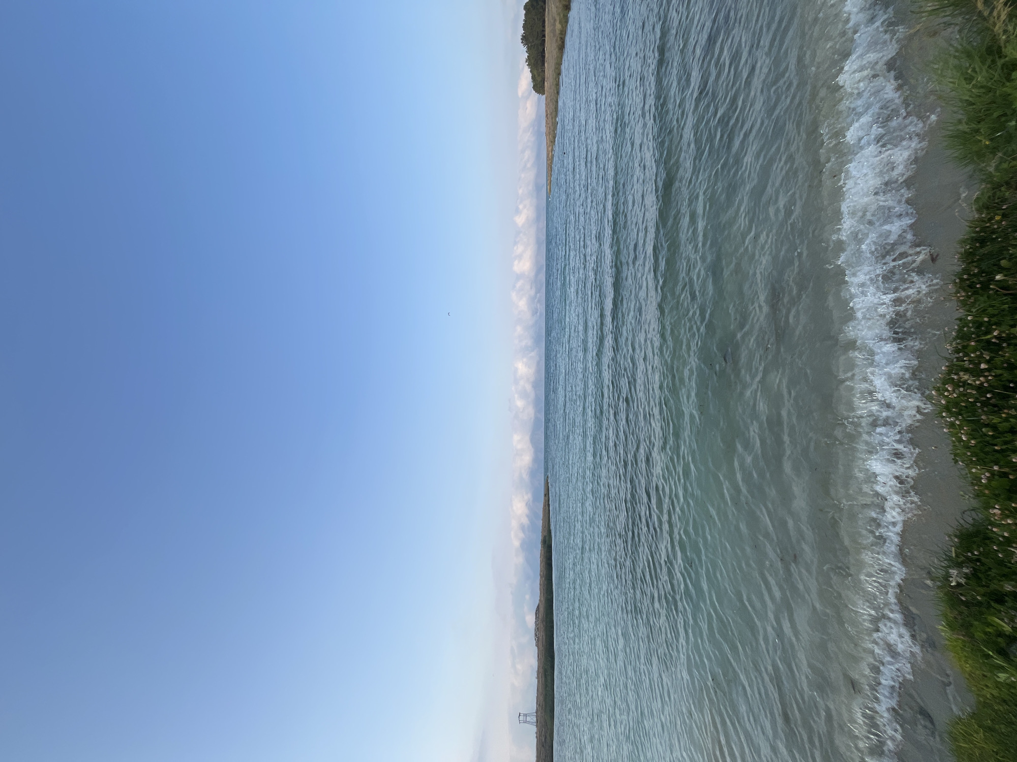 The image shows a tranquil body of water, likely a lake or a bay, with a clear blue sky above. Small waves gently lap at the sandy shore, which is lined with green grass and small white flowers. In the distance, a thin strip of land is visible, covered with vegetation and a structure resembling a watchtower. The water is a beautiful shade of turquoise and appears calm, reflecting the sky. The scene evokes a sense of peace and natural beauty. While the image is captivating, it does not contain enough information to determine its specific location within the city of Norashen, Armenia.