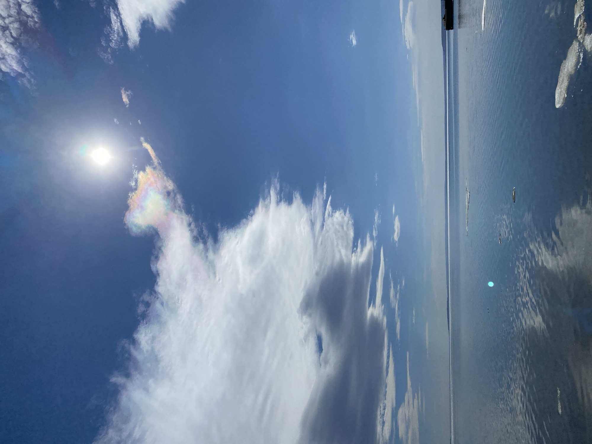 The image depicts a daytime view of the sky and water, likely a river or lake. The sky is predominantly blue with scattered clouds. Near the sun, there's a patch of iridescent cloud, showing a rainbow-like coloration. The water reflects the sky and clouds, with some ripples on the surface. Given the context, this could be the Volga River near Ulyanovsk, Russia. No people are visible in the frame. The focus is on the natural elements of sky and water, with an emphasis on the cloud formation. There are small icebergs on the water.