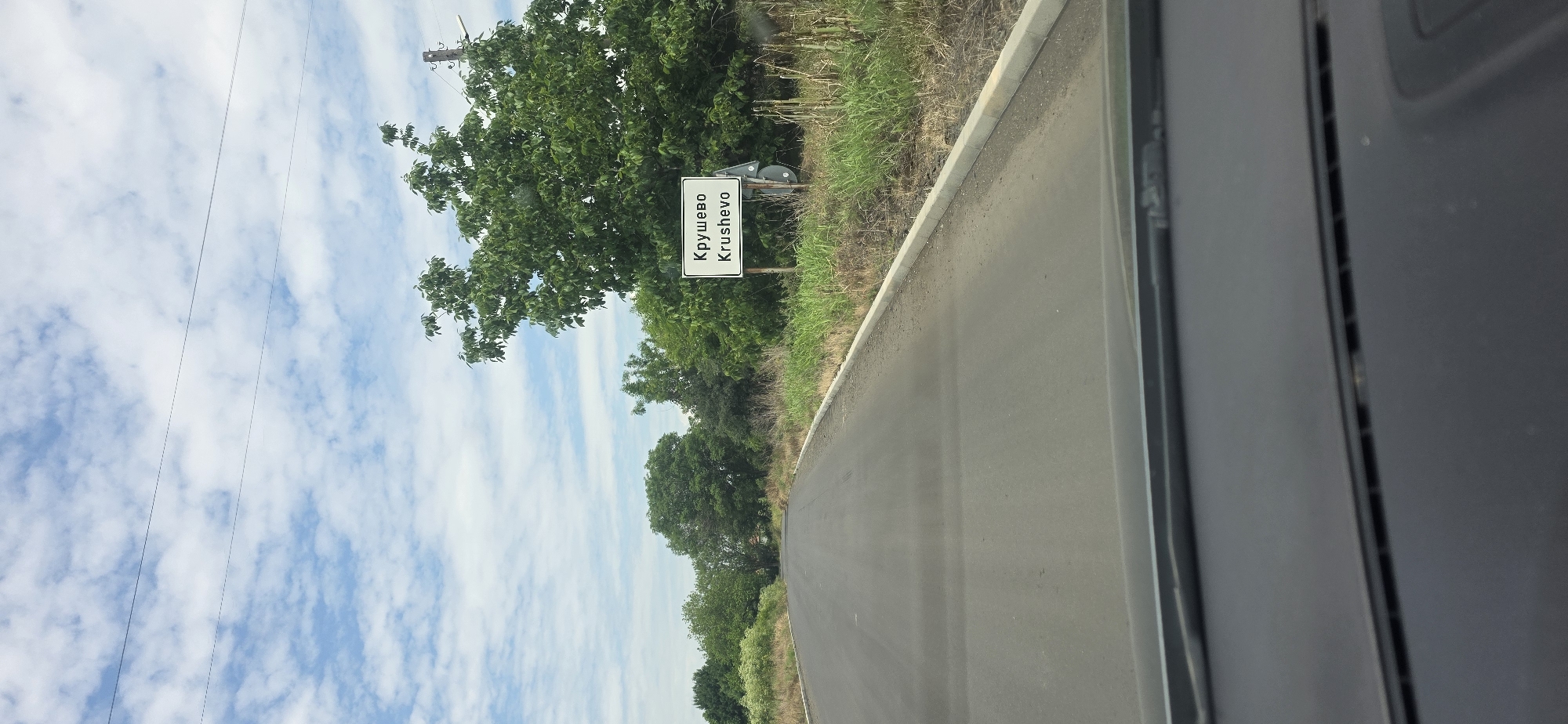 The image presents a forward-facing view from the interior of a vehicle, capturing an asphalt road leading into the distance. A white rectangular sign, supported by two dark posts, is prominently displayed on the right side of the road. The sign features text in black, identifying the location as "Крушево" (Cyrillic) and "Krushevo" (Latin script), indicating the entry point to the settlement of Krushevo, Bulgaria.
The road surface is dark asphalt, appearing dry, with a light-colored concrete curb delineating its right edge. Both sides of the road are flanked by dense green vegetation, primarily trees and tall grasses, which appear to be wild growth. The foliage is particularly thick on the right, partially obscuring the background behind the sign.
The sky occupies the upper portion of the frame, displaying a light blue color with numerous scattered white, fluffy clouds, indicative of daytime conditions. Two thin, dark utility wires are visible stretching horizontally across the upper sky. The dark-colored dashboard and the base of the windshield of the vehicle are visible in the lower foreground of the image. No individuals or animals are visible in the scene.