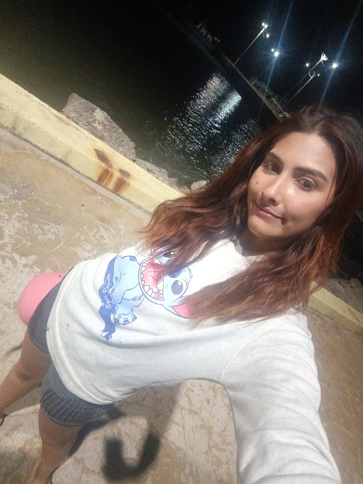An image taken at night in Juan Griego, Venezuela, depicts a woman positioned closely to the camera, likely self-photographed, with a waterfront background. The woman, with long, reddish-brown hair, is wearing a white long-sleeved shirt featuring a blue and pink animated "Stitch" character graphic. She has a faint smile and gazes directly into the camera. Her lower body, partially visible, includes dark shorts and a pink object to her left side. She appears to be leaning against or sitting on a light-colored, possibly concrete surface. The immediate foreground shows this textured concrete ground, with a yellowish concrete or wooden barrier along the left and top edges exhibiting dark, rust-colored staining. Beyond the barrier is a dark body of water reflecting multiple bright lights from the background. The background features several illuminated streetlights on poles, indicating an urban or dockside environment, with a dark landmass or structure visible across the water. The overall scene is illuminated by artificial light sources, casting some shadows.