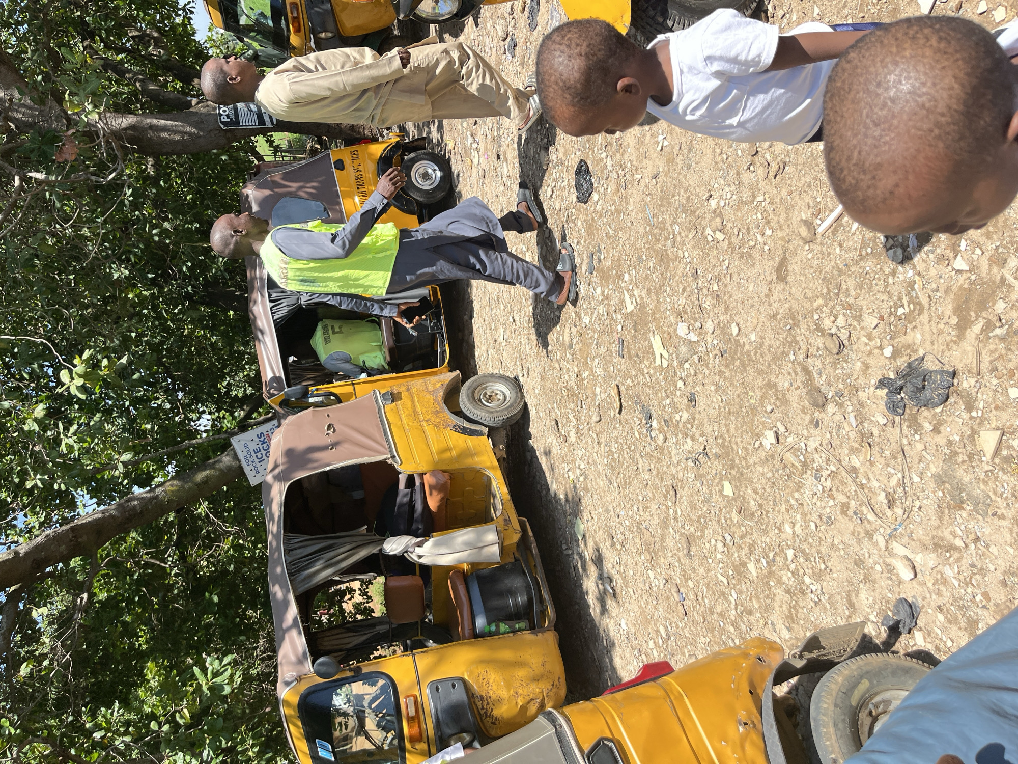The image captures an outdoor scene in Galadimawa Village, Nigeria, under bright daylight. The foreground consists of an uneven, unpaved dirt and gravel surface, scattered with small stones, dark plastic debris, and fragments of light-colored material.
In the midground, three yellow motorized rickshaws, commonly known as Keke Napeps, are present. The leftmost rickshaw features a brown soft top and a passenger compartment with brown seats and grey curtains tied back. Its yellow body shows signs of wear and multiple dents; a small green plastic bottle is visible on its floorboard. Slightly behind it, a second yellow rickshaw is visible, displaying "NDAU TRANS SERVICES" on its side, with another adult male wearing a neon yellow safety vest (marked with '5' on the back) seated inside. A third yellow rickshaw is partially visible on the far right.
Two adult males with dark complexions and short hair are walking from left to right across the midground. The lead male wears a grey long-sleeved shirt, a neon yellow safety vest, and brown and black open-toed sandals, holding a dark smartphone in his left hand. The second male, walking slightly behind and to his right, is dressed in a light tan long tunic and light-colored sandals.
In the right foreground, two young children with dark skin are visible. One child, wearing a white t-shirt, is positioned in profile, looking towards the right. The head of a second child is in the extreme foreground, partially obscuring the first child, also facing right.
The background is dominated by large green trees, with a thick tree trunk running diagonally upwards from the center. Two white signs with black text are attached to trees. One, above the leftmost rickshaw, partially reads "FOR ROCKSOLID ICE, ICE BLOCKS." Another, on a tree trunk near the middle rickshaw, shows "PO" at the top. Strong shadows are cast across the ground, indicating direct sunlight from above.