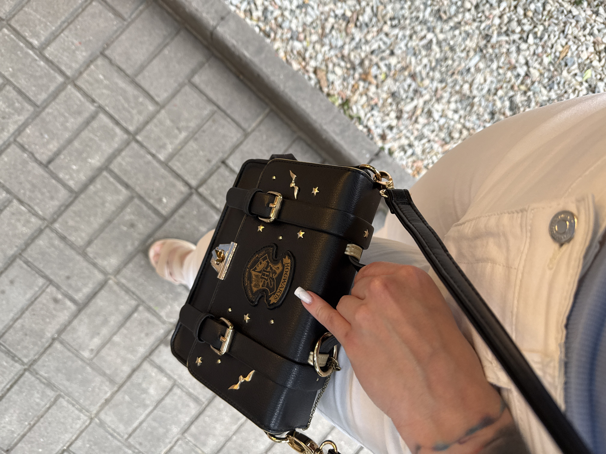 This is a medium-angle, eye-level shot of a black handbag decorated with gold stars and a lightning bolt. The bag has buckles and is attached to a strap. A hand with white nail polish is visible, touching the bag. The bag is positioned over the lap of a person wearing white pants and a light blue shirt. The person's feet are visible in the bottom left corner, wearing light-colored sandals.
The setting appears to be outdoors, with a paved area in the foreground made of grey rectangular tiles. In the background, there is gravel and a grey curb.
The emblem on the bag features the text "HOGWARTS" and other symbols, suggesting it is a themed item, possibly related to Harry Potter.
The lighting suggests it is daytime, with no clear indicators of weather conditions. The overall mood is casual and everyday. The location is identified as Almaty, Kazakhstan.