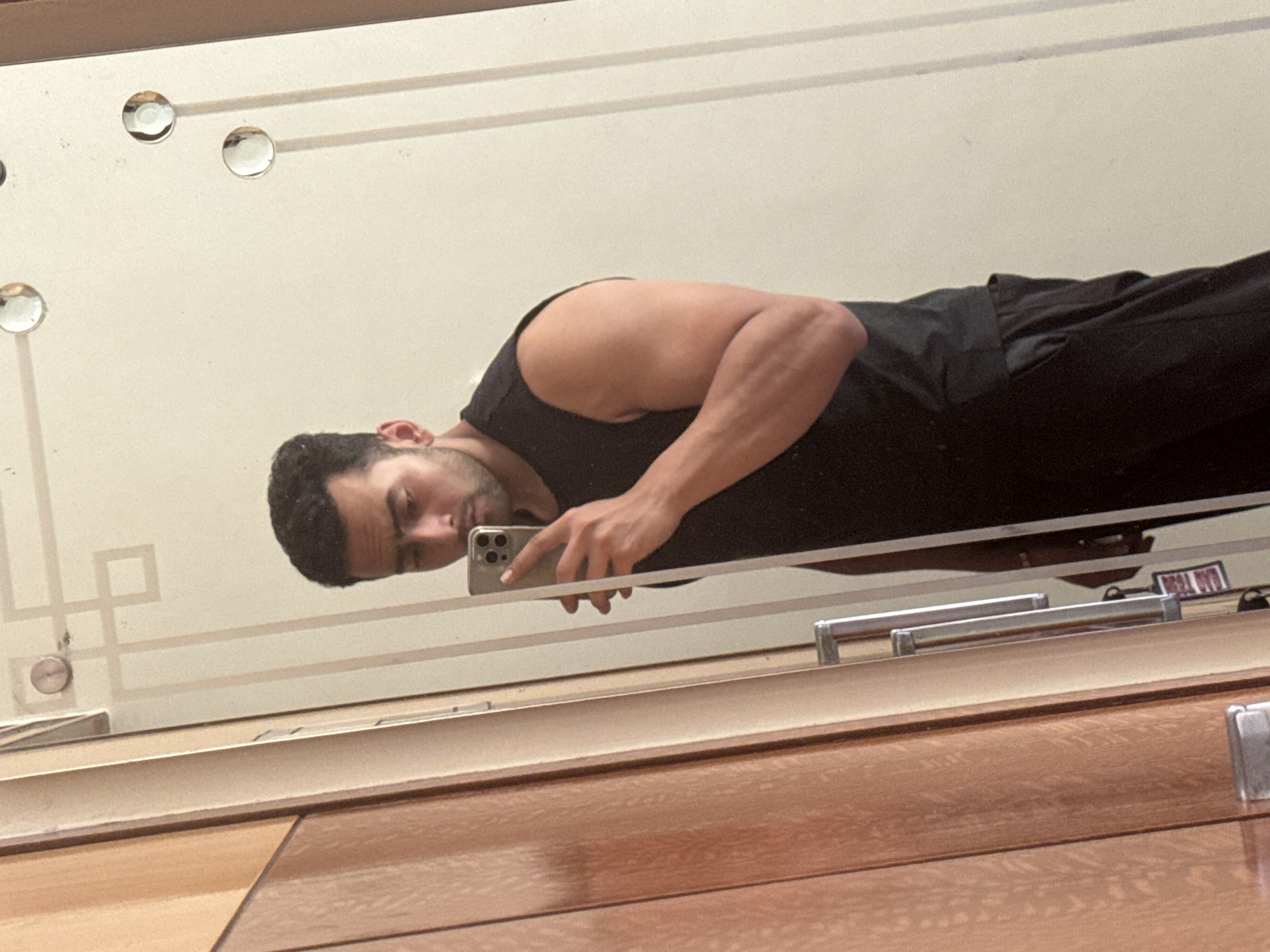 A man is taking a selfie in a mirror. The mirror has decorative elements, including circular accents and a geometric pattern. The man is wearing a black tank top and black pants. The background appears to be a plain wall. A dresser or cabinet is visible beneath the mirror. The location cannot be determined from the image.