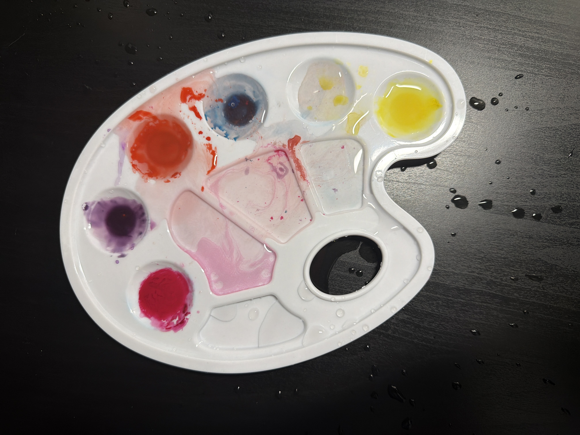A white, kidney-shaped plastic artist's palette is depicted, holding various watercolor paints mixed with water. It rests on a dark, textured surface, likely a tabletop, which is also sprinkled with numerous water droplets.
The palette features multiple circular wells and rectangular mixing areas. On the left side, a vibrant red and a deep purple paint are visible in circular wells. In the upper part, a dark blue and a light yellow paint occupy other wells. Several shades of pink and magenta are spread across the rectangular mixing areas, appearing diluted and swirled with water. The presence of water droplets on both the palette and the surrounding dark surface suggests recent artistic activity, possibly painting or cleaning the palette.
A prominent thumbhole is located on the right side of the palette. Near this thumbhole, faint, embossed text or numbers appear to read "50". The lighting is soft and even, suggesting an indoor setting. No people are visible, nor are there any discernible location cues within the image that would specifically indicate Moscow, Russia. The scene captures the aftermath of a creative session.