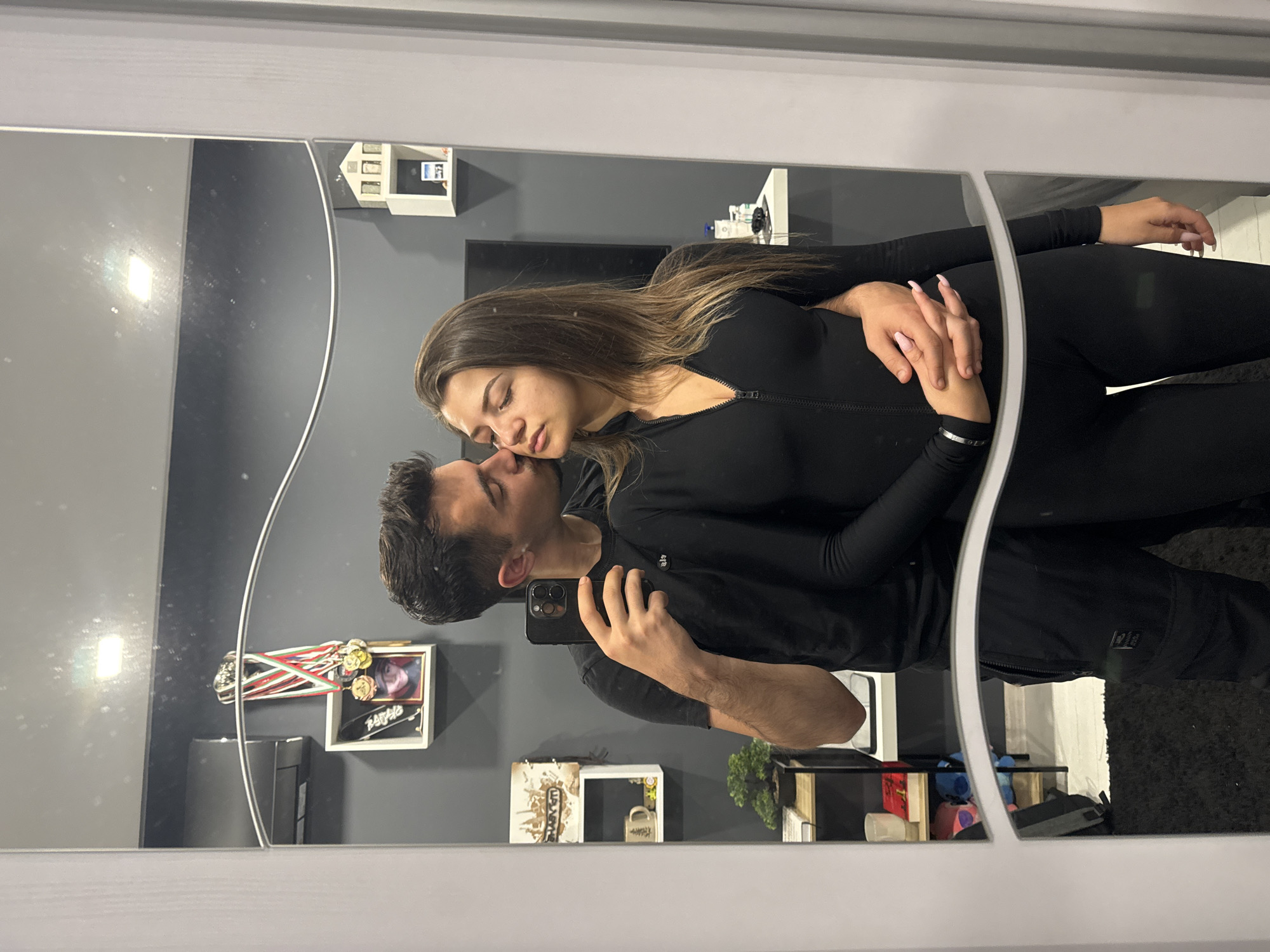 This media file is a mirror selfie featuring a young couple in an affectionate pose, captured indoors. The image is rotated 90 degrees clockwise, showing the subjects and their reflection turned on their side.
The woman, with long, straight brown hair, is wearing a black long-sleeved top, possibly a form-fitting jumpsuit or catsuit, with a zipper visible at the front. Her eyes are closed or partially closed as she leans into the man, resting her head near his cheek or neck. She has notable long, light pink fingernails. The man, with short dark hair and stubble, is taking the picture, holding a dark smartphone (likely an iPhone) in his right hand. He wears a black t-shirt, and his left arm is gently around the woman's waist. Their interaction conveys intimacy and tenderness.
The background, reflected in the large mirror with a white frame, reveals a modern interior space with dark gray walls. Various decorative items are displayed on white shelves. On the upper right, a miniature white house and a small framed item are visible. Behind the man's head, a shelf holds medals with red, white, and green ribbons, along with a framed picture and a black plaque bearing the text "PAYING". Another shelf towards the bottom left displays a white mug with a tree design and a small artificial plant. A television screen is discernible on the wall behind the woman. On a lower surface to the bottom right, colorful plush toys, including a blue character resembling Stitch, are present, and a dark-colored carpet is visible on the floor. A glare from an indoor light source is reflected in the top-left portion of the mirror.
The setting appears to be a home, potentially in Sofia, Bulgaria, based on the provided location context. The time of day is not indicated, but the scene is brightly lit by artificial light.