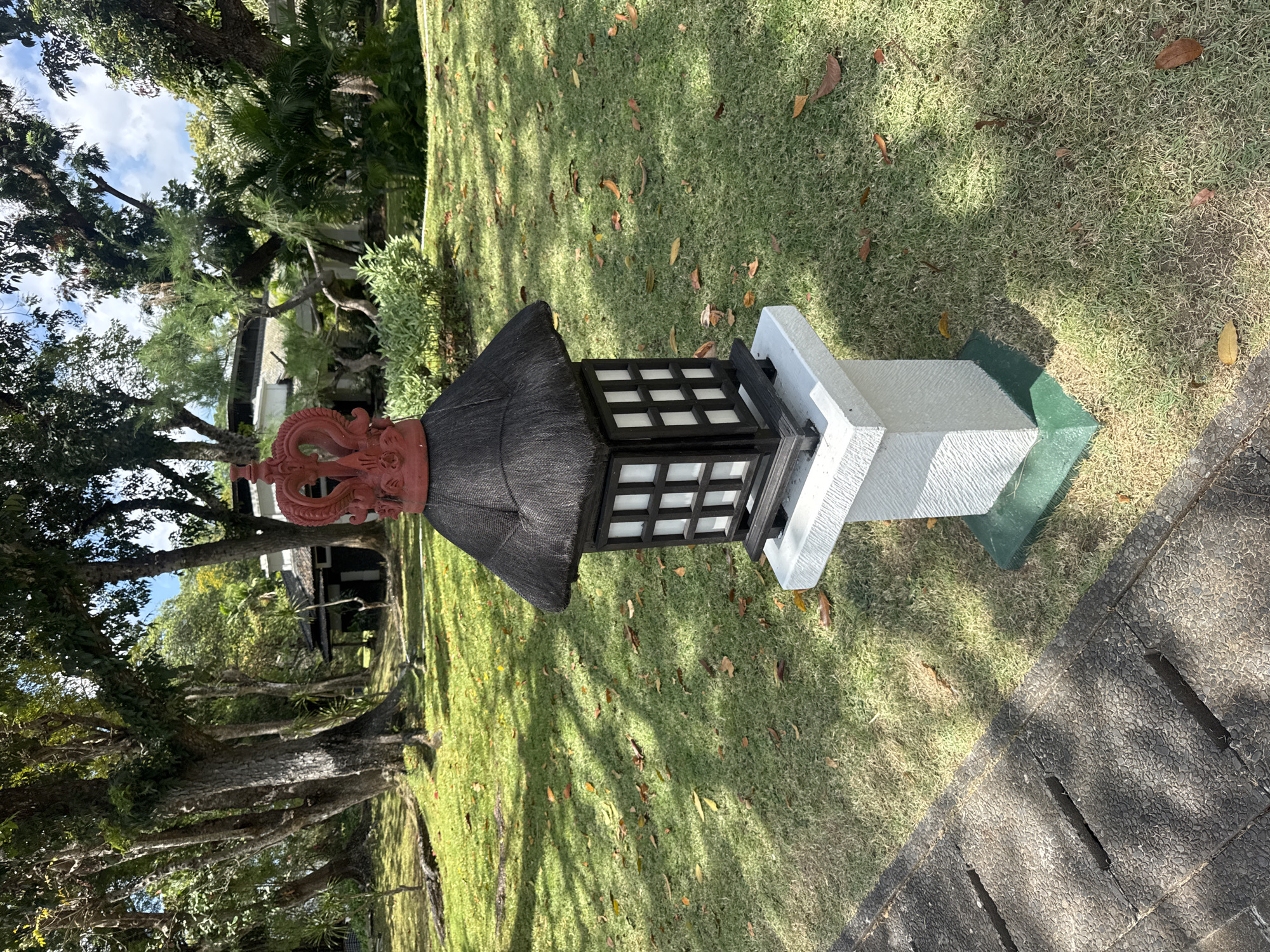 A medium shot captures a Japanese-style lantern in a grassy outdoor setting, likely a garden or park. The lantern, made of dark wood with white-paned windows, sits atop a white stone pedestal that is surrounded by a green base. The lantern is capped with a dark, thatched roof and is crowned with a red, ornate finial resembling a dragon or a crown. The grass surrounding the lantern is short and well-maintained, with scattered brown leaves on its surface. In the background, large trees with dense foliage create a natural canopy, casting dappled shadows on the grass. Beyond the trees, a building with a dark roof and white walls is visible, suggesting the presence of structures within the location. A stone pathway with a textured surface runs along the lower left corner of the image. The scene is well-lit, indicating daylight conditions. This image does not contain any people.