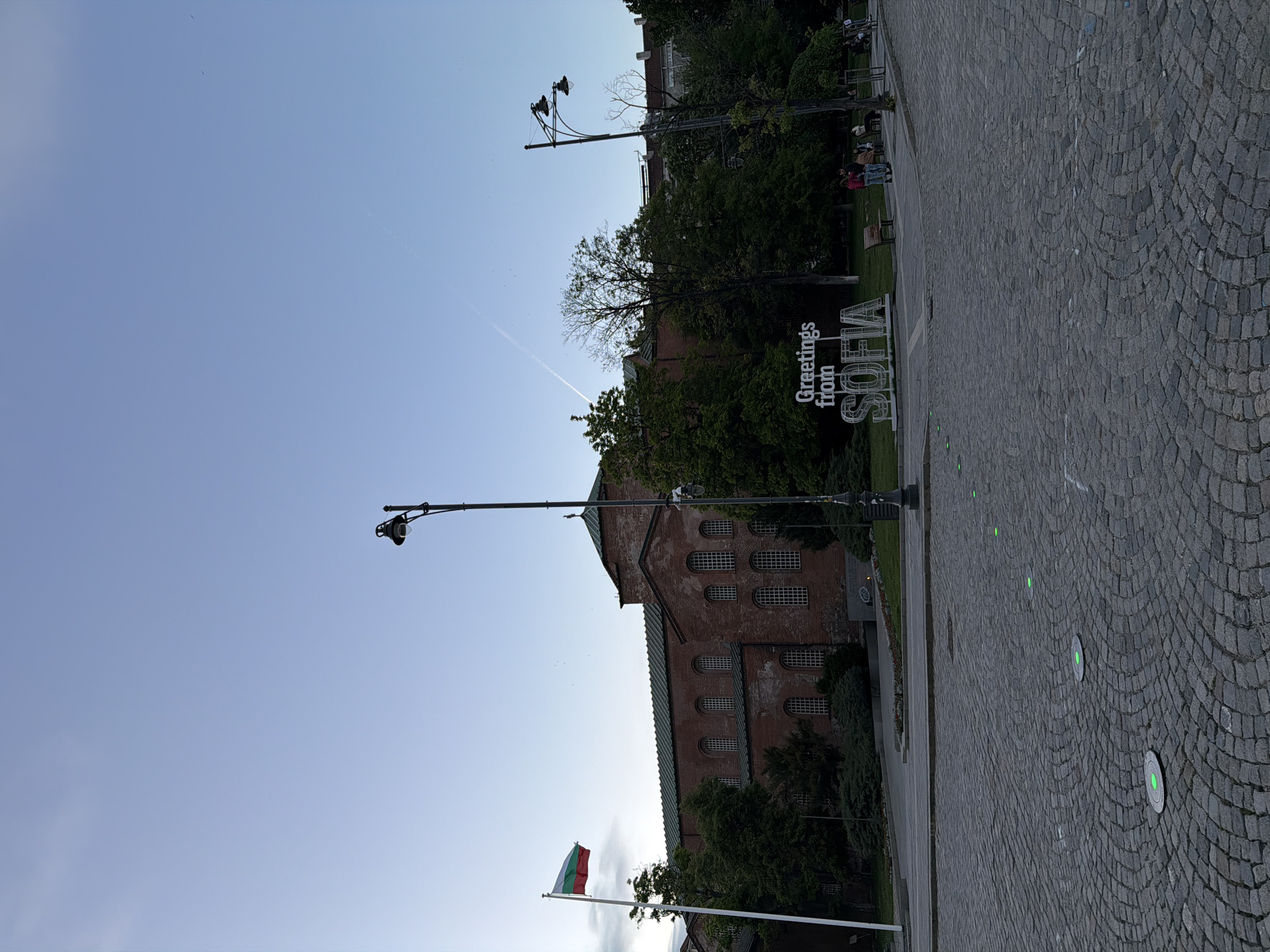 The image depicts a daytime scene in Sofia, Bulgaria. The foreground features a cobblestone path, leading towards a sign that reads "Greetings from SOFIA." Behind the sign, a historic brick building with multiple rows of windows is visible, partially obscured by trees. A Bulgarian flag is flown on a pole near the building. Streetlights are present, and a faint contrail is visible in the blue sky above. In the background, more buildings and trees can be seen. There is a small group of people in the far background.