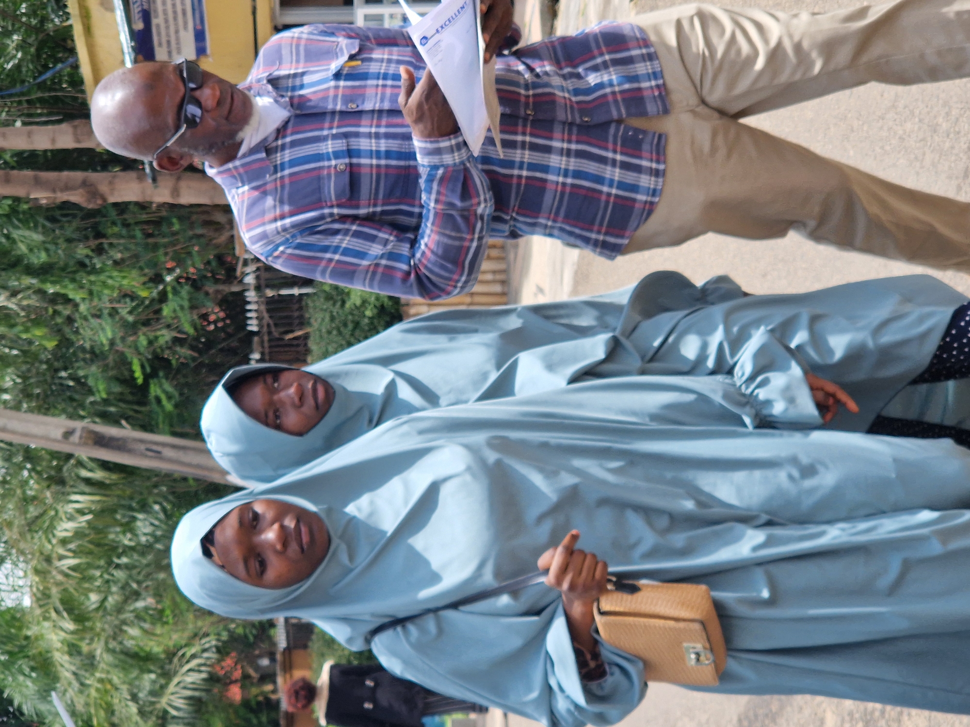The image depicts a street scene in Jos, Nigeria. In the foreground, two young women are walking, both wearing light blue hijabs and loose-fitting attire. The woman on the left carries a small, light brown woven bag on her left shoulder. To the right, a man wearing a blue and red plaid shirt, sunglasses, and khaki pants walks while looking down at a document he is holding. In the background, there are trees, a building with a yellow facade, and a power pole. The ground appears to be paved. The overall impression is of people walking outdoors in a moderately sunny environment.