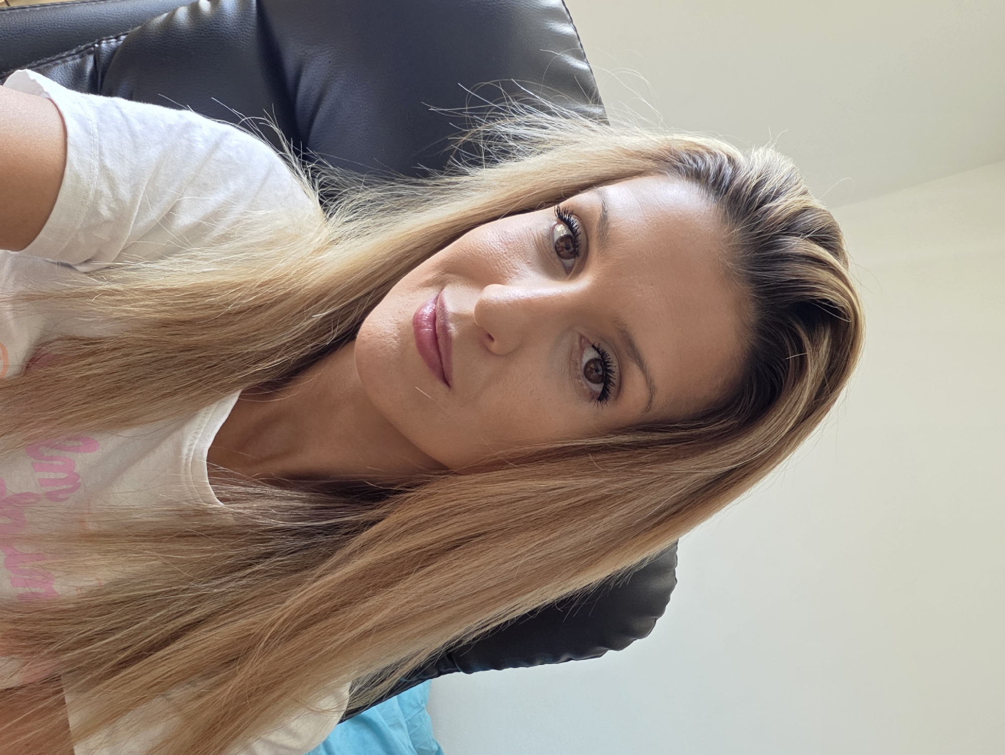 A woman with long, blonde, highlighted hair is seated in a black leather office chair in front of a white wall. She is wearing a white t-shirt with pink lettering. Her face is turned towards the camera, and she is looking directly at the viewer. She has brown eyes, dark eyelashes, and pink lipstick. The lighting suggests an indoor environment with natural light coming from the left. The background is plain and out of focus. There is a hint of a blue object, possibly a pillow or blanket, on the right side of the image. The specific location in Munich, Germany cannot be determined from the image.