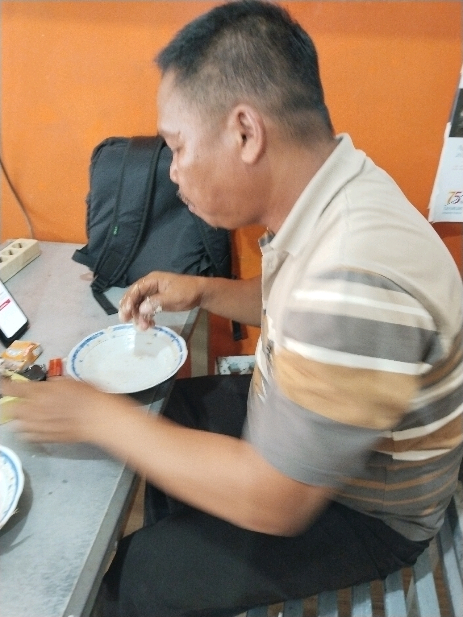 A man with short black hair, wearing a beige and grey striped polo shirt and black pants, sits at a table in what appears to be an indoor establishment. He is actively engaged in eating from a white plate with a blue rim, using his hands. A black backpack is placed behind him on the table. In front of him, also on the table, are a mobile phone and some small packaged items. The environment features an orange wall in the background and a table with a grey surface. This scene is located in Prigi, Indonesia.