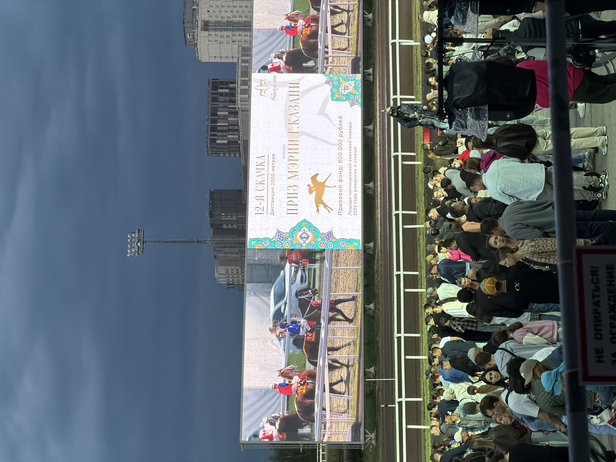 A panoramic view of a horse racing event is displayed on a large outdoor screen, while a crowd of spectators gathers below. The screen shows a horse race in progress, with jockeys on horseback and a car in the background. Text overlays on the screen provide race details, including "12-Я СКАЧКА," "Дистанция 2000 метров," and "ПРИЗ МЭРИИ Г.КАЗАНИ." The crowd, composed of diverse individuals, stands and watches the screen, with some holding phones to take pictures. The environment appears to be a racecourse with a track and fencing. The sky above is dark and cloudy, suggesting a possibility of rain. There is a person in a distinctive outfit standing in the foreground, possibly an entertainer or staff member. In the background, modern buildings and a tall lamppost with floodlights are visible.