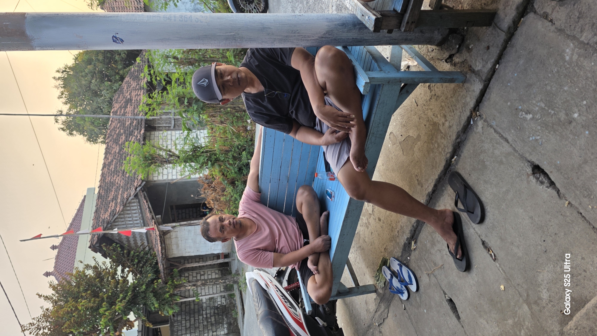 Two adult males are seated on a light blue wooden bench outdoors in Juwana, Indonesia. The man on the left wears a pink t-shirt and black shorts and is seated with his legs crossed. The man on the right wears a black t-shirt, grey shorts, and a black baseball cap with a white logo. He is also seated with his legs crossed. A small plastic bottle and a packet of cigarettes are on the bench between them. A motorcycle is parked to the left of the bench. The background shows a residential building with a tiled roof and a tree. The ground is paved concrete with cracks. Two pairs of flip-flops are on the ground near the bench.