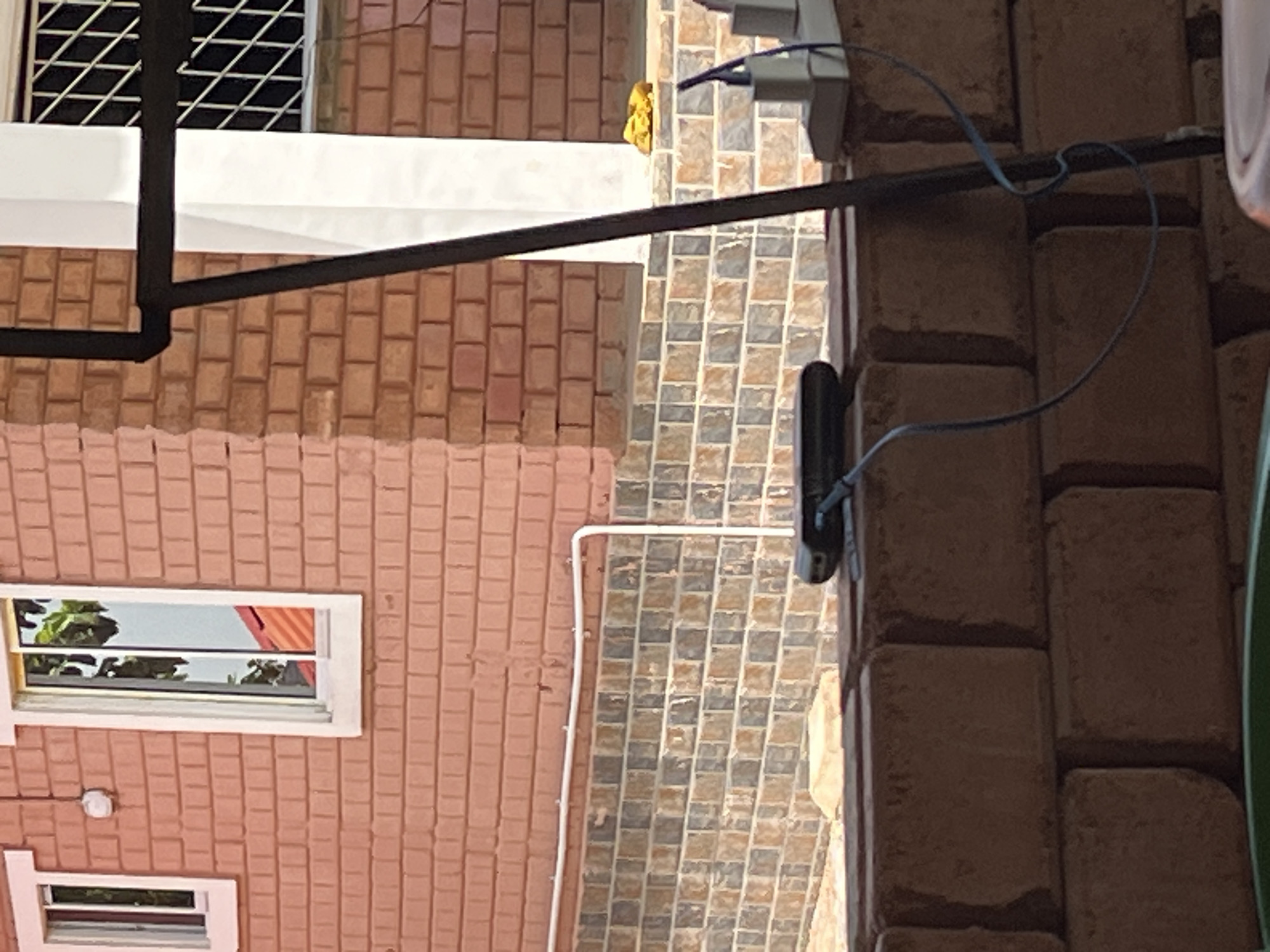 The image shows a close-up view of the exterior of a building under construction, likely in Jos, Nigeria. The building features pink brick walls with a decorative tiled lower section. Two windows are visible: one is a simple rectangular window with a white frame, and the other is a larger sliding window with a white frame, reflecting greenery and a red-tiled roof. A white pipe runs horizontally along the wall above the tiled section. In the foreground, a dark electronic device, possibly a portable charger, is connected to a blue cable, resting on a low brick wall. To the right, a white electrical outlet with two plugs is also visible, with a cable extending from it. The overall impression is of an unfinished building, with construction materials and electrical accessories present.