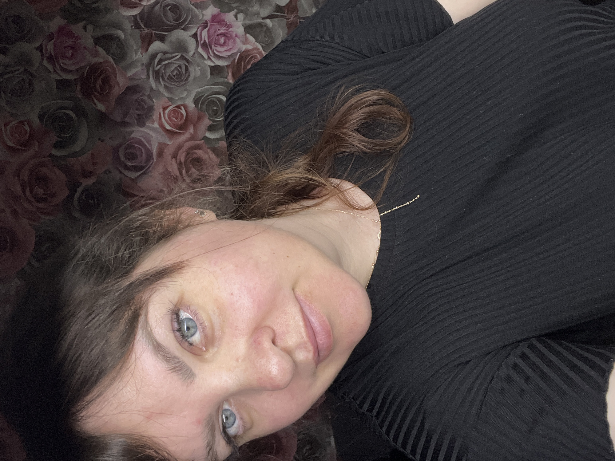 A woman is lying down, facing the camera. She has fair skin, blue eyes, and brown hair. She is wearing a black, horizontally-ribbed shirt and a thin gold necklace. The background consists of a floral wallpaper pattern with roses in shades of red, pink, gray, and white. There is no evidence to suggest this picture was taken in Noviy, Russia.