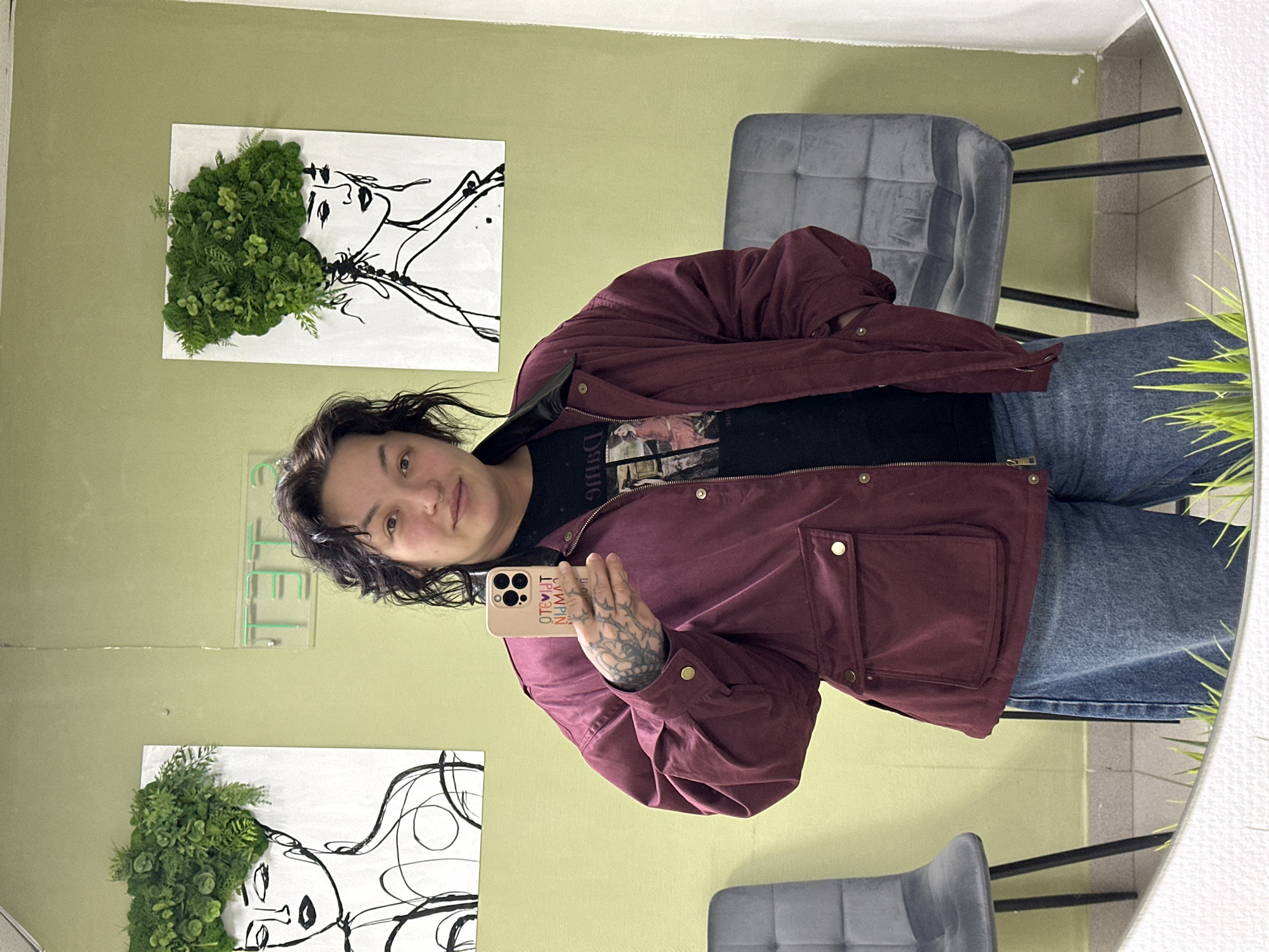 A person is taking a selfie in a mirror inside a room. The individual is wearing a dark-colored t-shirt, a maroon-colored jacket, and blue jeans. They are holding a phone with a light-colored case. The room has light green walls, decorated with two pieces of art featuring drawings of faces with plant-like hair. Two gray chairs are also visible. Based on visual clues, this photo was taken indoors but no specific location in the city of Chaikovsky, Russia, is discernible.
