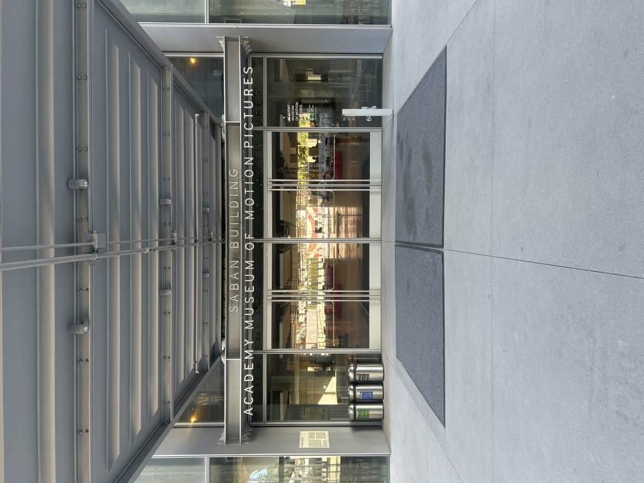 The entrance to the Saban Building Academy Museum of Motion Pictures is shown. Gray metal bars and a corrugated roof overhang the glass doors. Inside, reflections of the museum's interior are visible. The gray tiled floor extends to the entrance with two gray mats just outside the doors. A set of trash and recycling bins is near the entryway.
