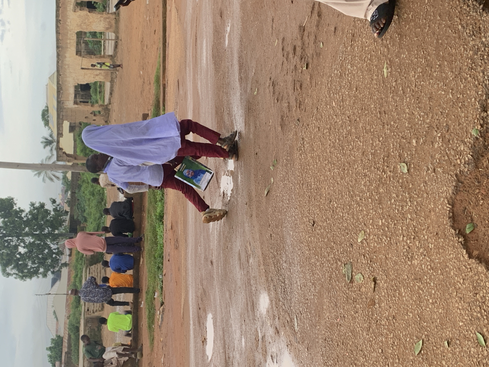 In the city of Bukuru, Nigeria, two children, one wearing a white shirt and maroon trousers and the other a white hijab and maroon trousers, walk across a muddy, gravelly dirt road. The child in front is carrying a framed picture of an elderly person. They are walking towards the right of the frame, away from a group of people sitting on a low wall in the background. The group consists of at least seven males, some in casual wear, and one individual in a pink hoodie. Behind them, partially ruined structures with open doorways and windows suggest a derelict building. A tall, thin tree stands to the left of the structures, and a palm tree is visible further in the background. The sky is overcast.