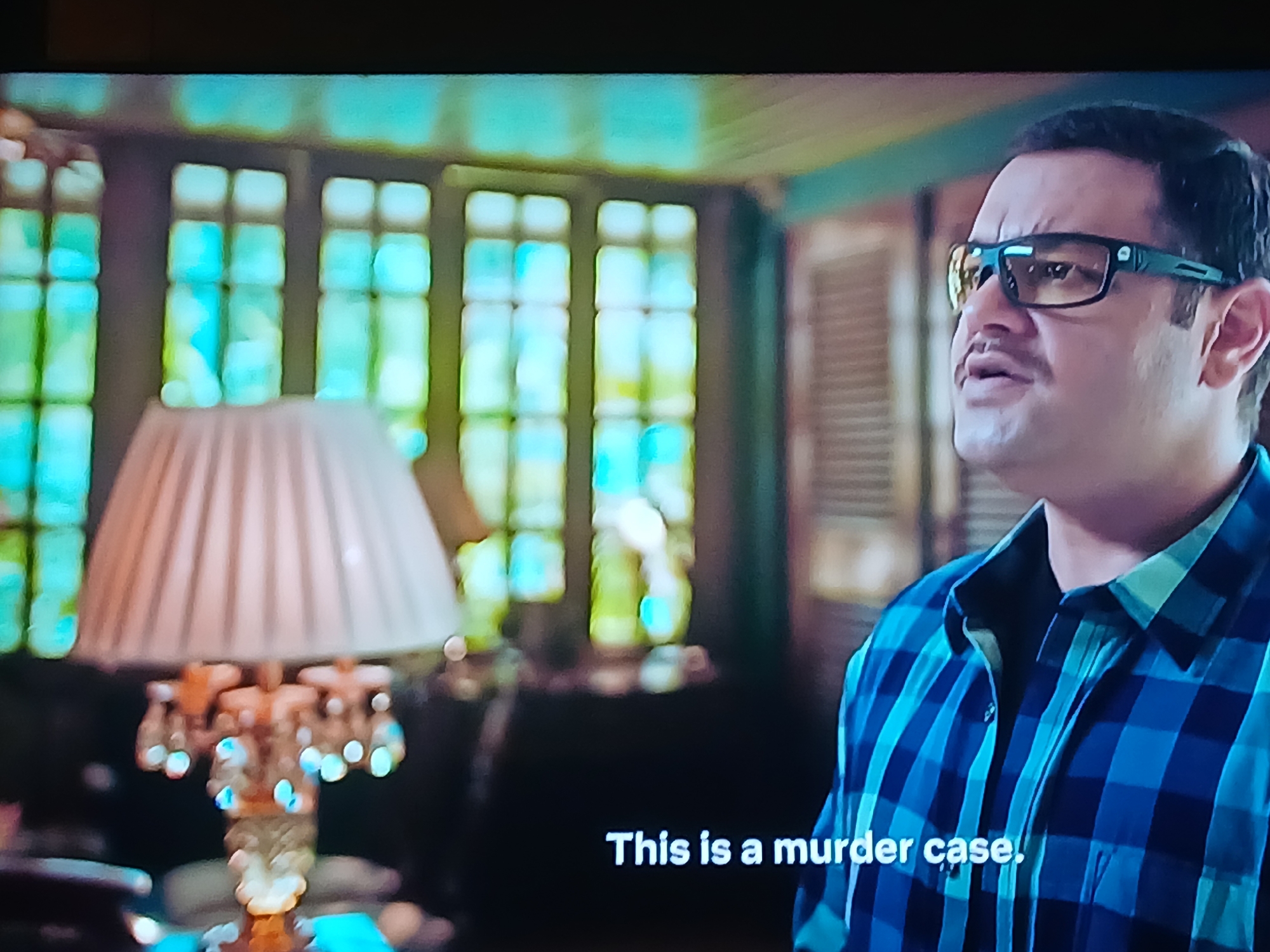 The image depicts a scene likely from a film or television show, with a man in the foreground wearing a blue and black plaid shirt and glasses. His facial expression appears serious. The text "This is a murder case" is superimposed at the bottom of the frame, indicating the context of the scene.
Behind the man, there is a lamp with a white shade and an ornate base. In the background, there are large windows with multiple panes, suggesting an interior setting.
It's impossible to determine the specific location in Bengaluru, India, based solely on this image. The scene's details (window design, lamp style, interior décor) could be found in various homes or buildings within the city. Without additional information, such as distinctive landmarks or architectural styles unique to a particular area of Bengaluru, it is not possible to place this scene geographically.