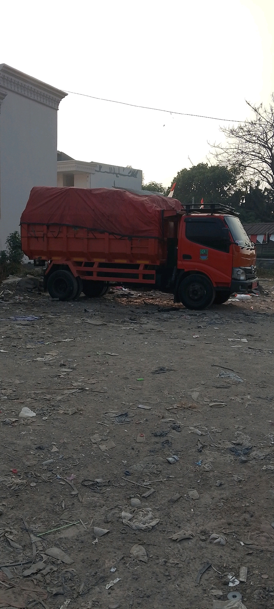 An orange truck is parked on a dirt lot littered with debris. The truck has a red tarp covering its cargo. In the background, there is a building with decorative trim and some trees. The sky is overcast.