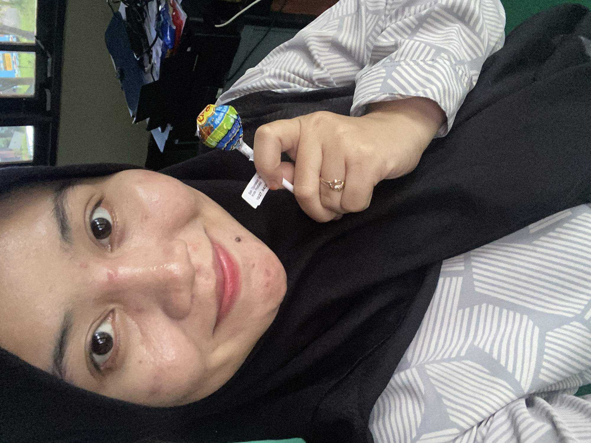 A medium shot shows a woman wearing a black hijab and a light-colored patterned shirt. She is holding a lollipop in her right hand, which is adorned with a gold ring. The woman is looking towards the camera and smiling. Her face has some blemishes. The background appears to be an indoor setting, possibly an office or study, with a desk and some objects on it. The overall lighting is natural and soft. The image seems to be a selfie taken by the woman.
