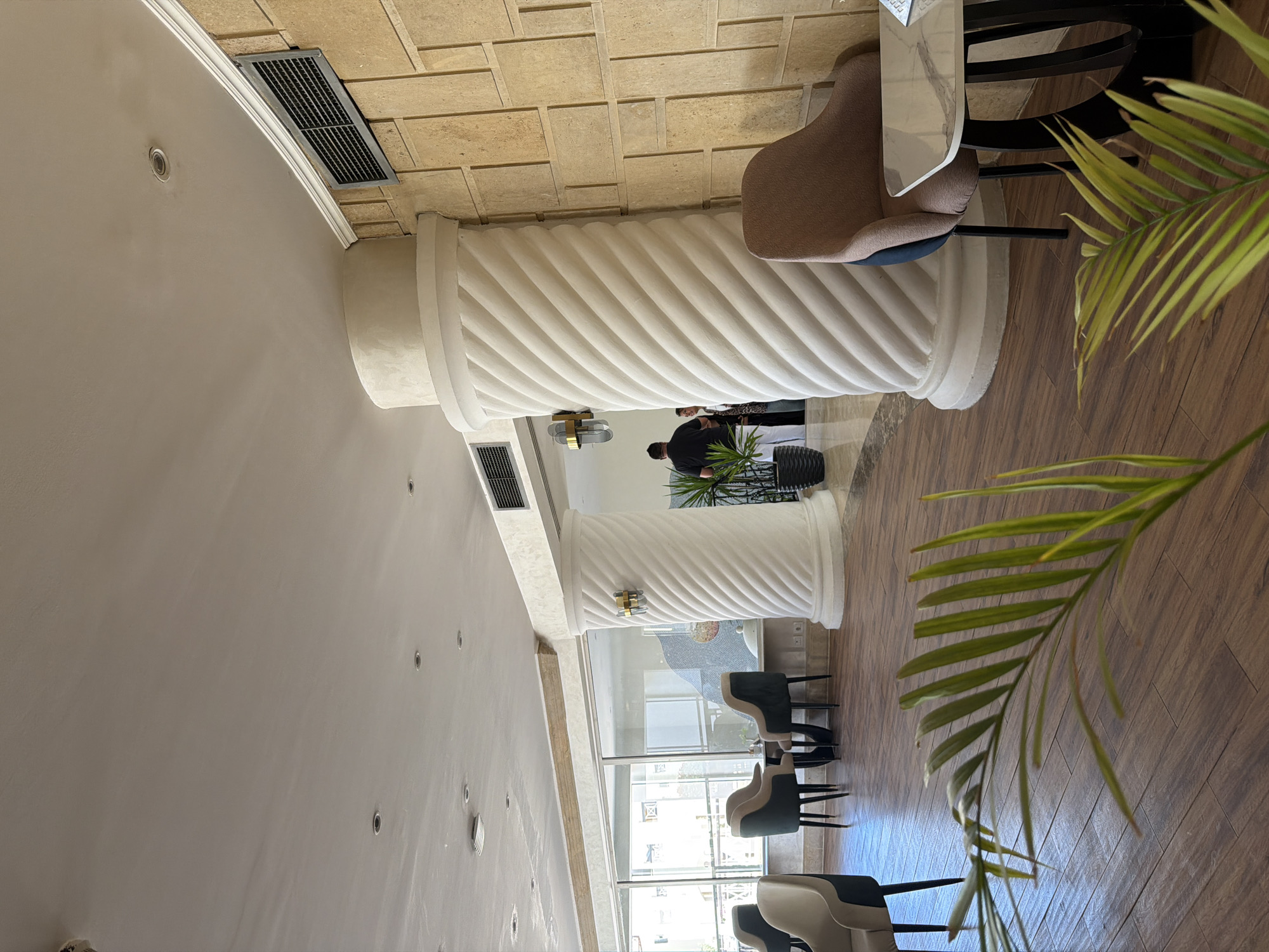 This is a photograph of the interior of a hotel lobby or lounge area in Sharm El Sheikh, Egypt. The scene features several elements of a hotel's common space. In the foreground, a large, tropical plant's fronds are visible, partially obscuring the view.
The middle ground shows a seating area with several upholstered chairs and small tables. Two chairs are grouped around a small round table. Another chair, a muted brown color with a dark blue accent, is situated next to a small marble-topped table. The chairs appear to be modern and comfortable.
Dominating the background are large, fluted decorative columns, one of which is particularly prominent and features a spiral design. Beyond the columns, large windows offer a glimpse of the exterior, suggesting a bright, possibly sunny day. The wall behind the columns is textured with a pattern resembling stacked stone or tiles in a tan or beige color.
There are two individuals visible in the background. One person, wearing a dark shirt and light pants, is standing near a potted plant. Another person, in the distance, is partially visible and appears to be interacting with someone else.
The environment is well-lit, with recessed ceiling lights and decorative wall sconces. The flooring is a polished wood-look laminate. The overall impression is one of a comfortable and contemporary hotel lobby, likely designed to be a relaxing space for guests.