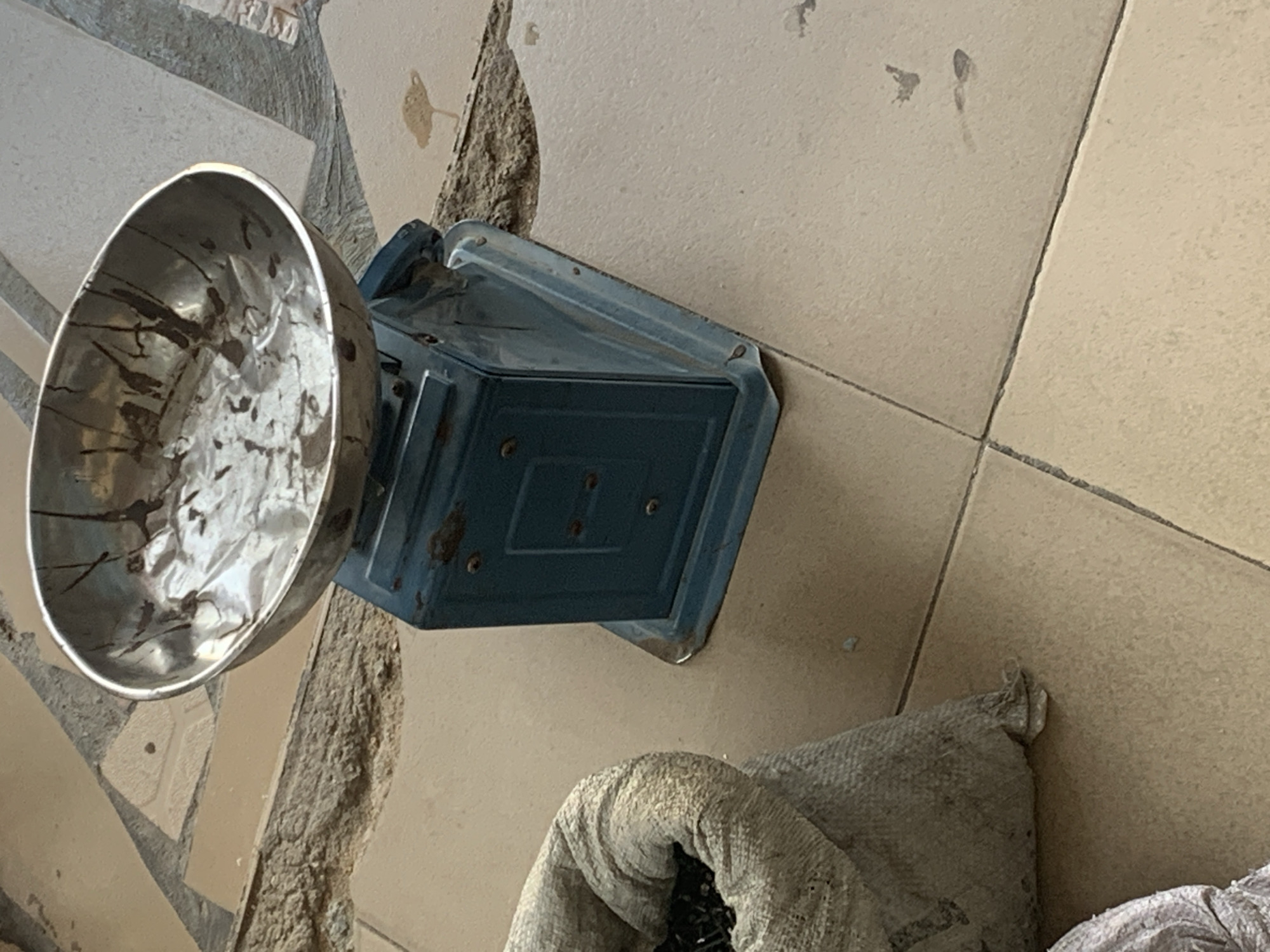 A blue metal scale sits on a tiled floor next to a burlap sack. The scale has a tarnished metal bowl on top, with dark streaks inside. The burlap sack is open, revealing dark contents, possibly beans or seeds. The floor is made of beige tiles with some dirt and debris visible. The background shows a rough, sandy surface and more tiles. There are no people or other animals present in the image. The overall impression is one of an outdoor or rustic setting, possibly a market stall or storage area.