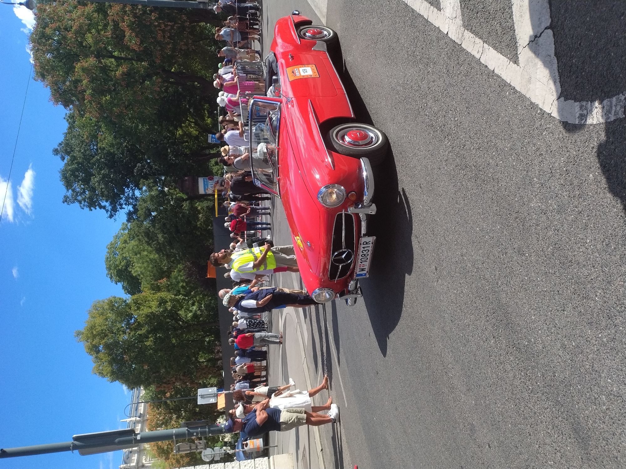 In Vienna, Austria, on a sunny day, a bright red vintage Mercedes-Benz convertible is parked on an asphalt street. The car, a 190 SL, has a black convertible top up and a white interior. The license plate reads "W 10831R". A driver in sunglasses and a white cap is visible in the driver's seat. The car has a sticker with the number "129" on the passenger side.
The street is lined with people, some standing behind a metal barrier, watching the car. Among the spectators are a man in a navy blue shirt and khaki shorts, a woman in a white dress, and a person wearing a yellow vest. In the background, there are trees and buildings, including a light-colored classical building with columns. The sky is blue with scattered white clouds. The scene appears to be part of a public event or parade.