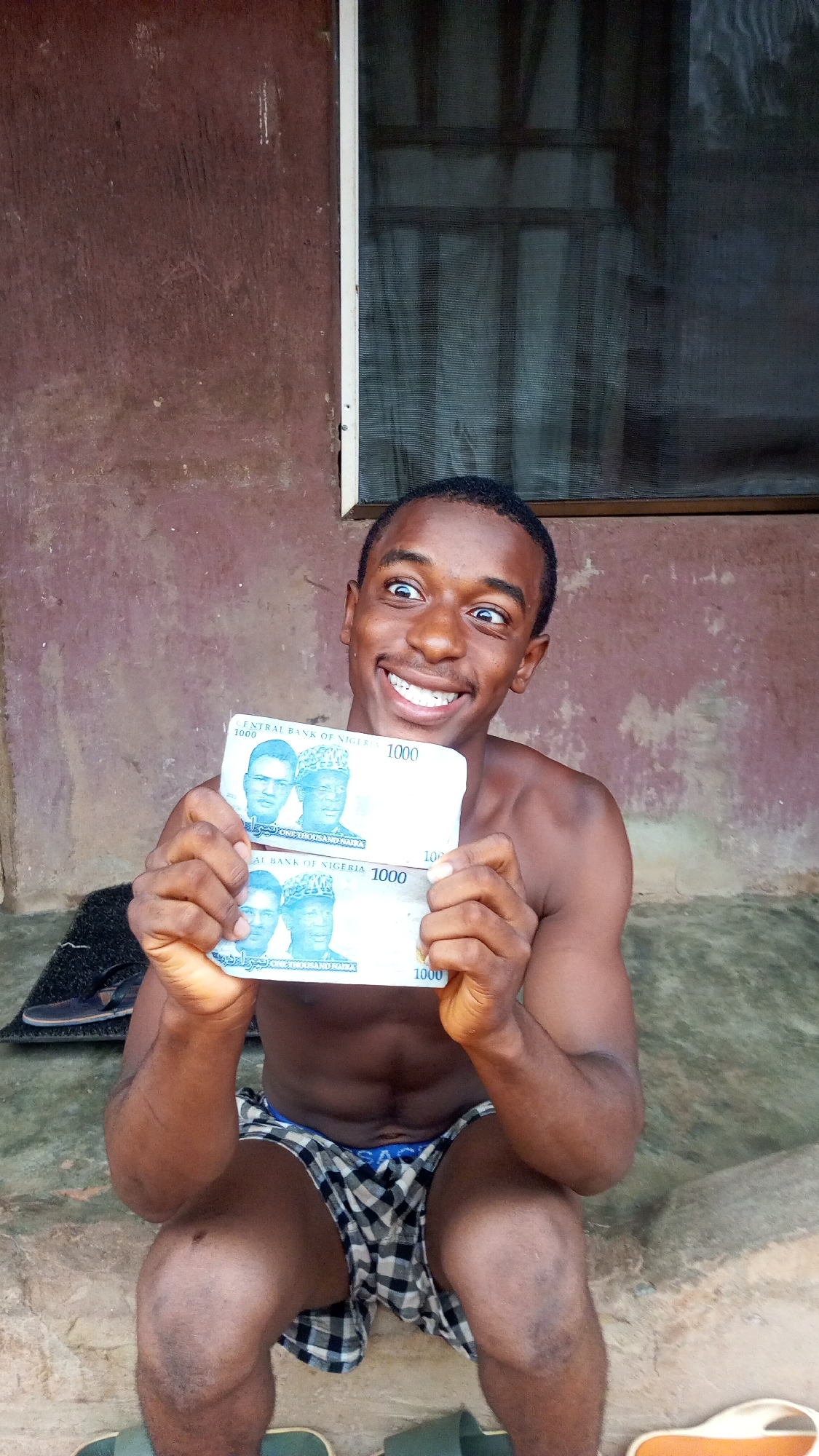A young, dark-skinned man, likely in his late teens or early twenties, is captured in a seated or crouching position, shirtless and wearing checkered boxer shorts. He holds two Nigerian 1000 Naira banknotes directly towards the camera with both hands. His face displays an exaggerated expression of joy and excitement, with wide-open eyes looking slightly upward and a broad, toothy smile.
The banknotes are clearly visible, featuring the text "CENTRAL BANK OF NIGERIA," the denomination "1000," and "ONE THOUSAND NAIRA," along with portraits of figures and Arabic script. The backdrop consists of a weathered, reddish-brown wall, suggesting an outdoor or partially sheltered setting. To the man's right, a window with a mesh screen and a white frame is visible. The ground beneath him appears to be a light-colored, hard surface like concrete. Near his feet, various sandals or shoes are visible: a dark pair on the left and light green and light-colored sandals on the right. The scene is illuminated by daylight, and given the context, it takes place in Onitsha, Nigeria. The man's animated expression conveys a strong sense of happiness or pride related to the money.