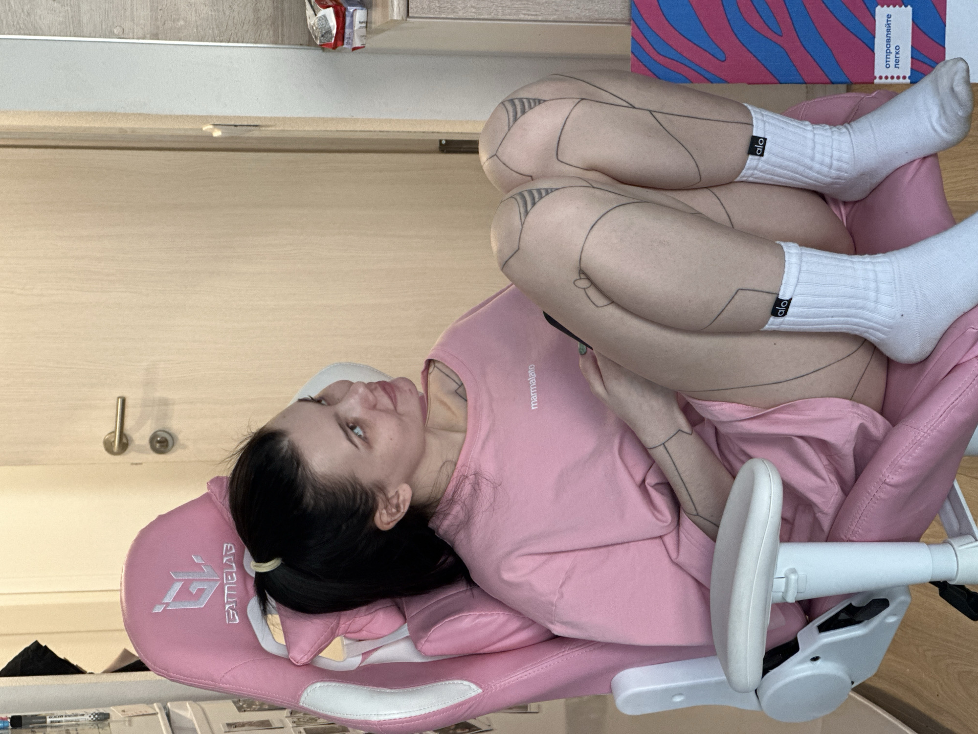 A fair-skinned woman with dark hair pulled back with a hair tie is seated in a pink gaming chair. She is wearing a pink t-shirt with the word "marmalato" printed on it. She is in an interior space, evidenced by the doorknob and frame in the background. Her legs are crossed, and she is wearing white socks with the "alo" logo. The woman has tattoos on her legs and arms. A box with blue and pink patterns is located behind the legs and bears a label with Cyrillic characters, which reads "отправляйте легко" which translates to "send easily". The chair has white armrests and the "GAMELAB" logo embroidered on its headrest. The location is Lyubertsy, Russia, based on the Cyrillic text. A cluttered desk is visible at the bottom of the frame.