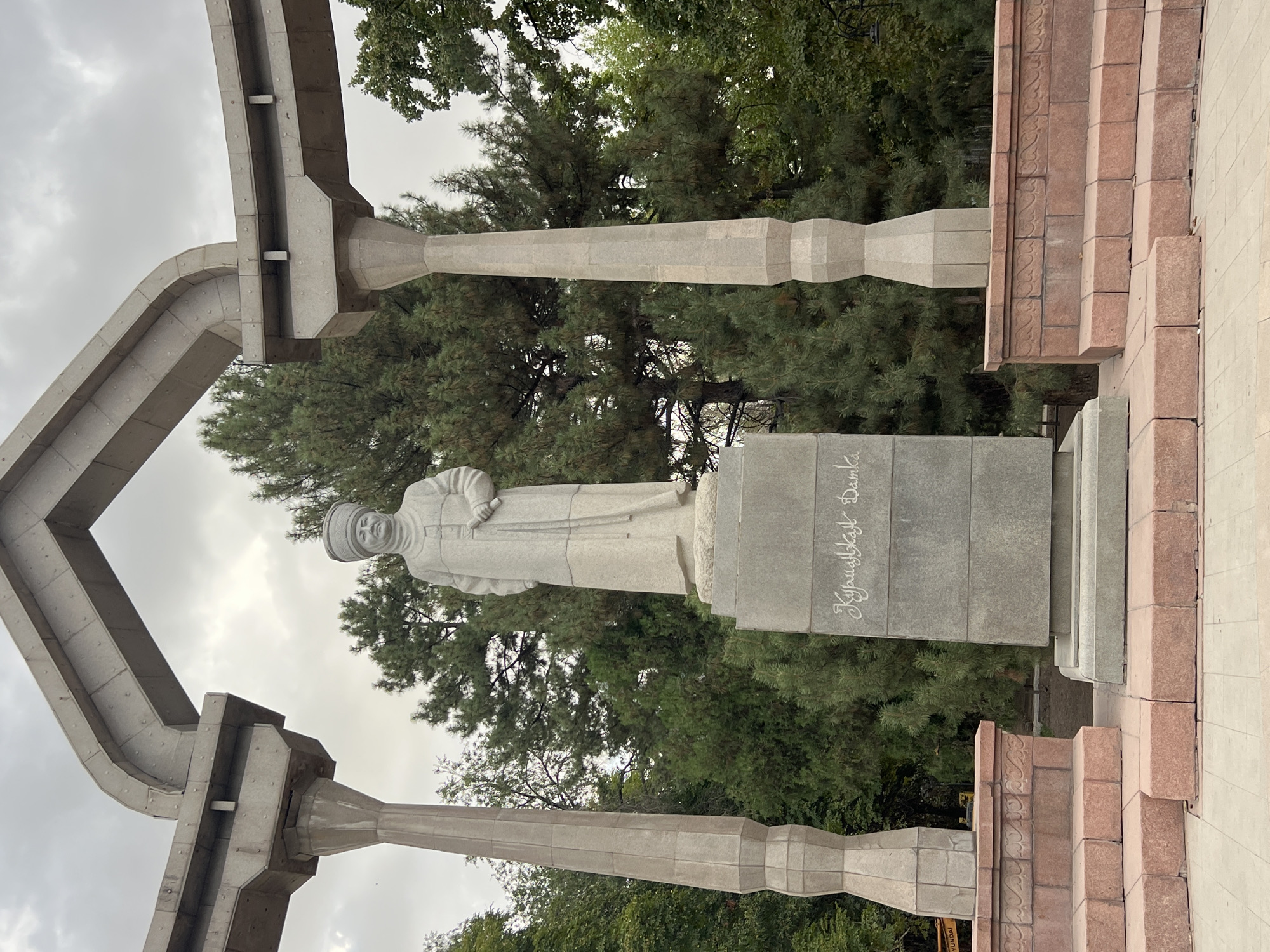 A stone statue of a man in traditional attire stands on a pedestal in Bishkek City, Kyrgyzstan, beneath a cloudy sky. The statue is positioned within a circular stone structure supported by four large, angular columns. The man in the statue wears a distinctive cap and a long robe, and he holds a scroll in his right hand. The pedestal is inscribed with what appears to be Cyrillic script. The surrounding area is characterized by lush green trees, primarily pines and deciduous varieties, and a paved walkway. The overall scene conveys a sense of historical monumentality.