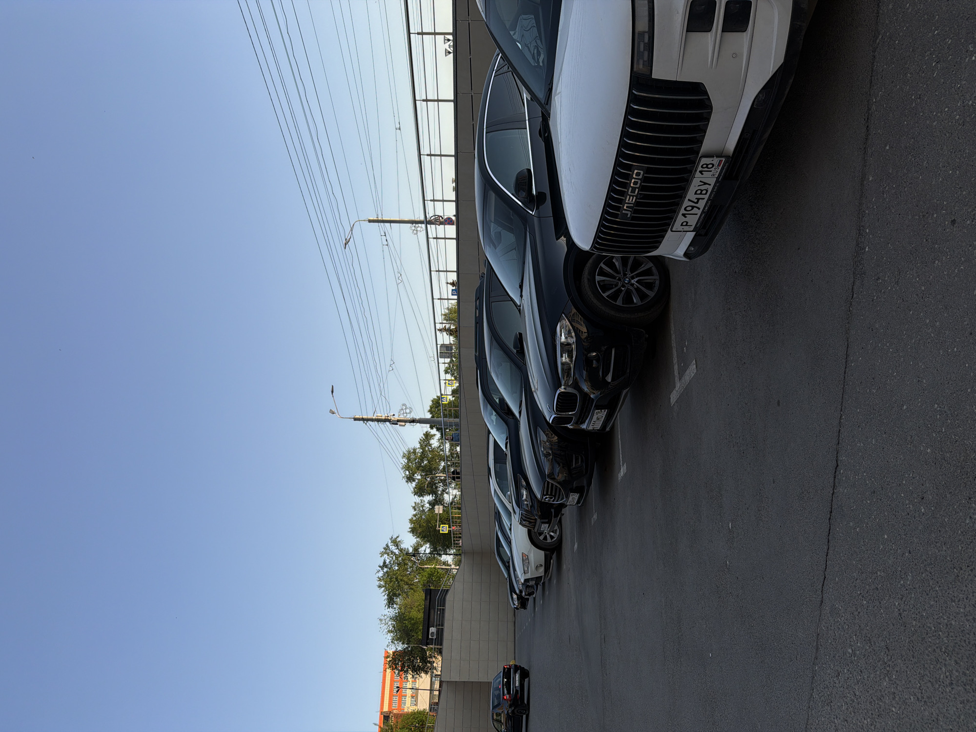 The image depicts a row of parked cars in Izhevsk, Russia. The closest car to the viewer is white with a prominent grill and a license plate reading "P194BY 18," indicating Russian registration. Behind it are several dark-colored cars. Overhead, power lines stretch across the sky. A building with a gray exterior is visible in the background, along with sparse greenery. In the lower left corner, an orange building is partially visible.