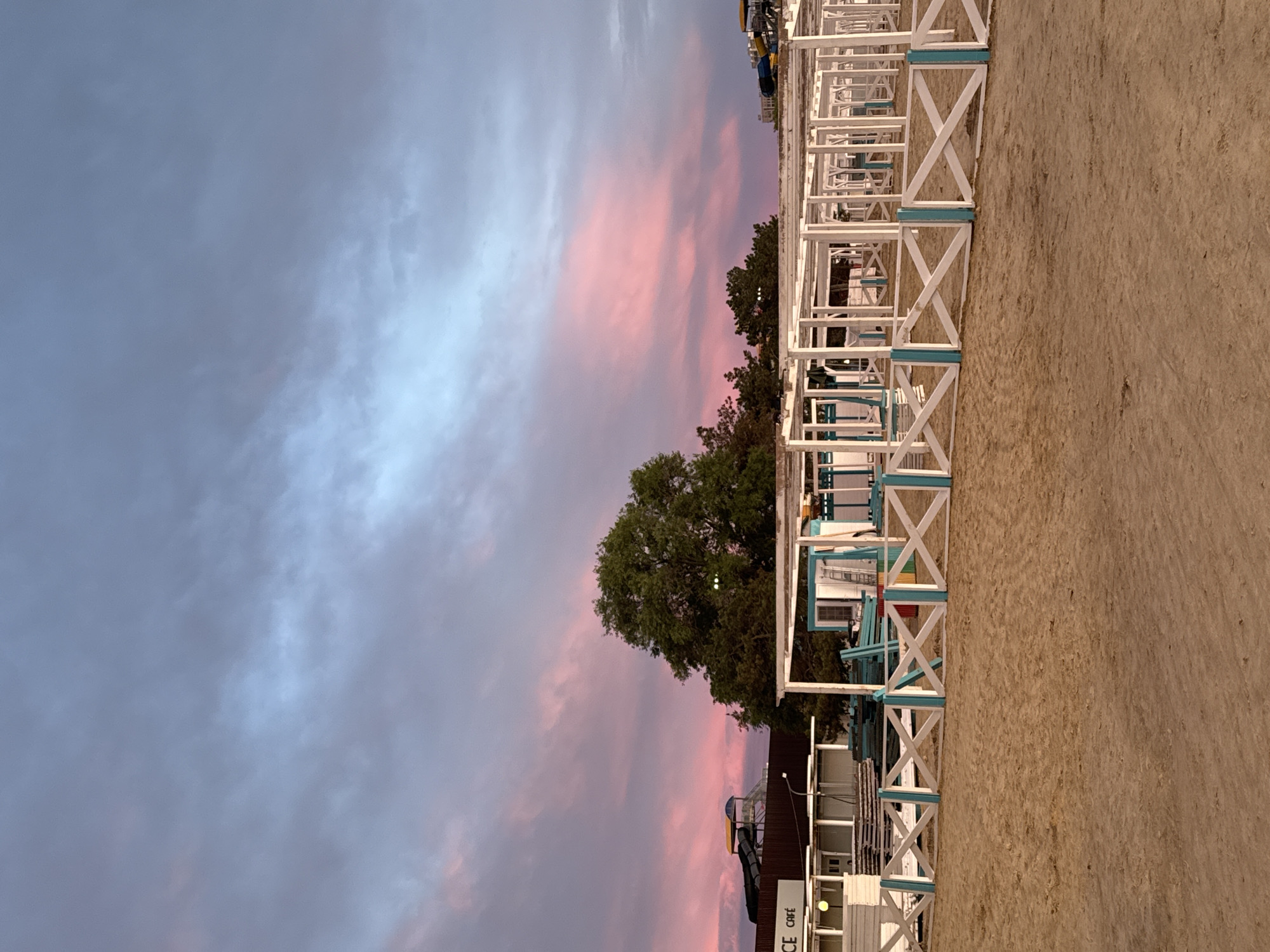 This media file captures a serene outdoor scene, likely a beach or waterfront area in Anapa, Russia, during either sunrise or sunset. The sky dominates the upper and left portions of the image, showcasing a beautiful blend of soft blue-grey clouds on the far left, transitioning into striking pink and orange hues towards the center and right, characteristic of twilight.
The foreground and bottom right feature an expansive sandy ground, typical of a beach. Along the right side, a series of identical white, open-frame structures, possibly beach cabanas or stalls, are arranged in a row. These structures have distinctive light blue crossbeam accents, and some appear to contain seating or other amenities, though no people are visible.
In the mid-ground, a large, lush green tree stands prominently, its foliage breaking the line between the sandy area and the colorful sky. Behind the tree and further along the right, other indistinct buildings and structures are partially visible, including what appears to be a section of a water slide. A partial sign "CE Café" (likely "CAFE" or "CAFÉ") is visible on one of the structures in the lower left. The overall atmosphere is peaceful and calm, with no activity or events explicitly taking place.