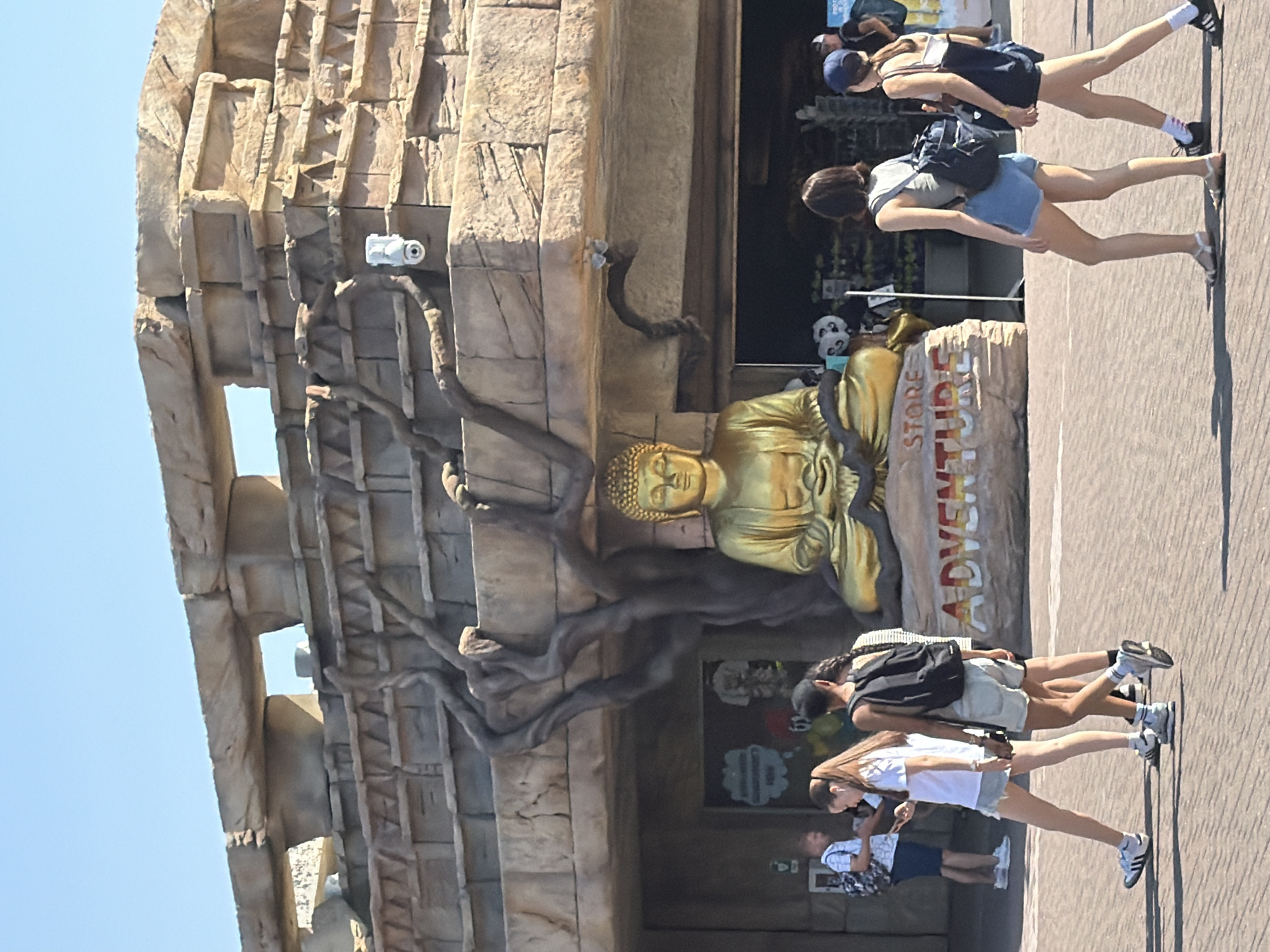 A golden statue of Buddha sits on a rock formation in front of a building with a stone-like facade and large, gnarled tree roots extending from it. The building appears to be a store, with a sign that reads "STORE ADVENTURE." The sky is clear and blue. In the foreground, several people are walking on a paved area. A man in a white checkered shirt and dark shorts is on the left, looking at his phone. To his right, a young woman in a white t-shirt and denim shorts walks past, also looking at her phone. Behind her, a person with a backpack walks with another person. On the right side of the image, two young women walk away from the camera. One wears a grey tank top and denim shorts, with a backpack. The other wears a white tank top and dark shorts, with a backpack. The scene is set in a sunny outdoor environment, possibly an amusement park or tourist attraction, given the "Adventure Store" sign and the general aesthetic. The architecture suggests a theme park or a resort. The location is identified as being in the city of Castelnuovo del Garda, Italy.