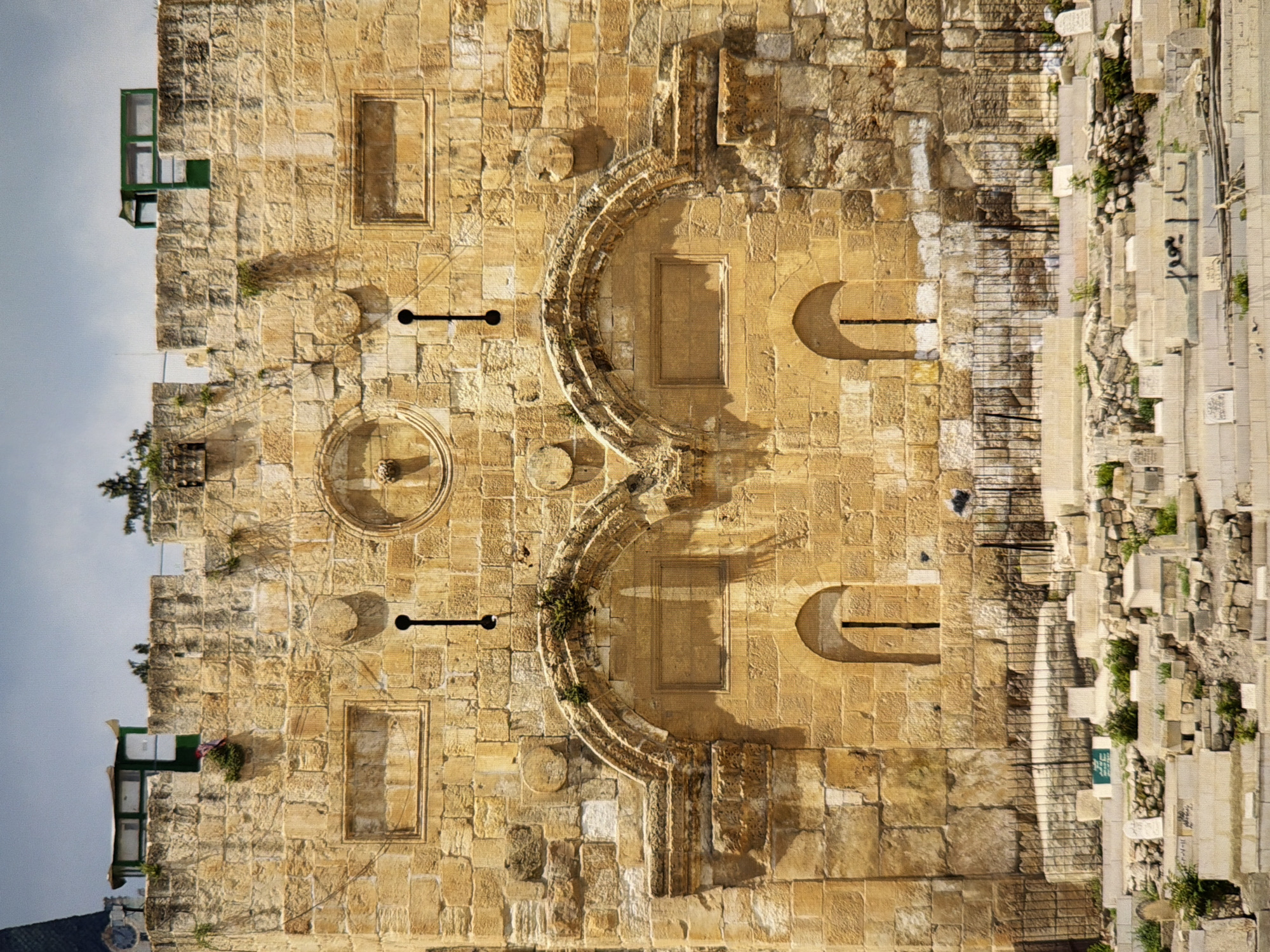 The image shows the Golden Gate of Jerusalem, an ancient stone gateway in the Old City walls. The gate is a monumental structure with two arched entrances, each featuring a small, arched window above. The stonework is weathered and a testament to its age. Above the arches, rectangular inscriptions are visible, and in the center, a circular relief is present. The top of the wall has crenellations and small green structures. In front of the gate, a cemetery is visible, with numerous gravestones and low fencing. The sky is overcast. The location is the Old City of Jerusalem, not Kyiv, Ukraine.