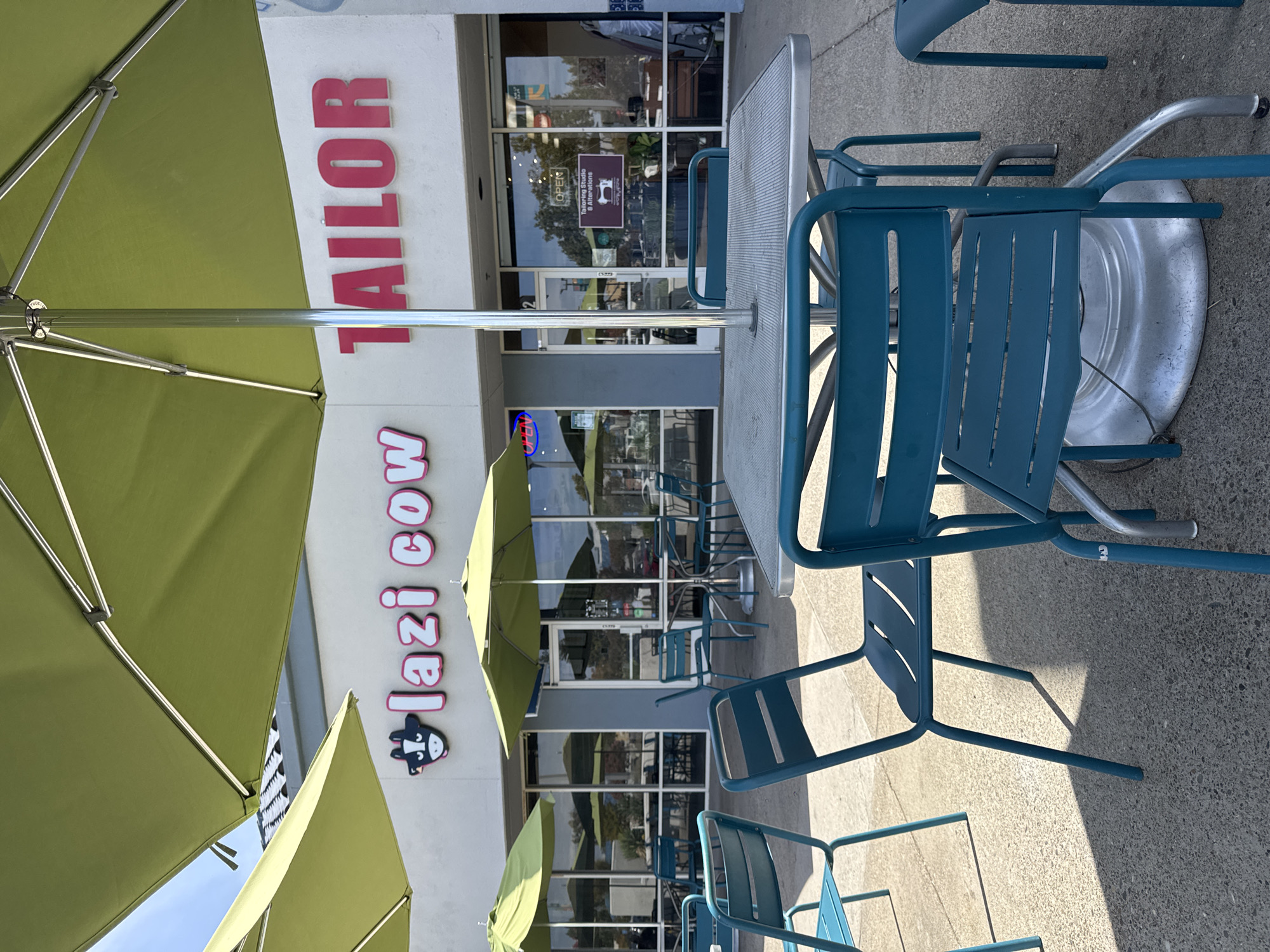 An outdoor seating area in front of a business features teal metal chairs and tables under large, lime green umbrellas. The building facade behind the seating area has a white wall with the pink neon sign "lazi cow" on the left and "TAILOR" in large pink block letters on the right. The windows of the building are large and reflective, showing interior views. One window displays an "OPEN" sign and a sign for "Tailoring Studio & Alterations". The ground is paved with light grey concrete. The scene is well-lit by natural daylight, casting shadows from the umbrellas and furniture.