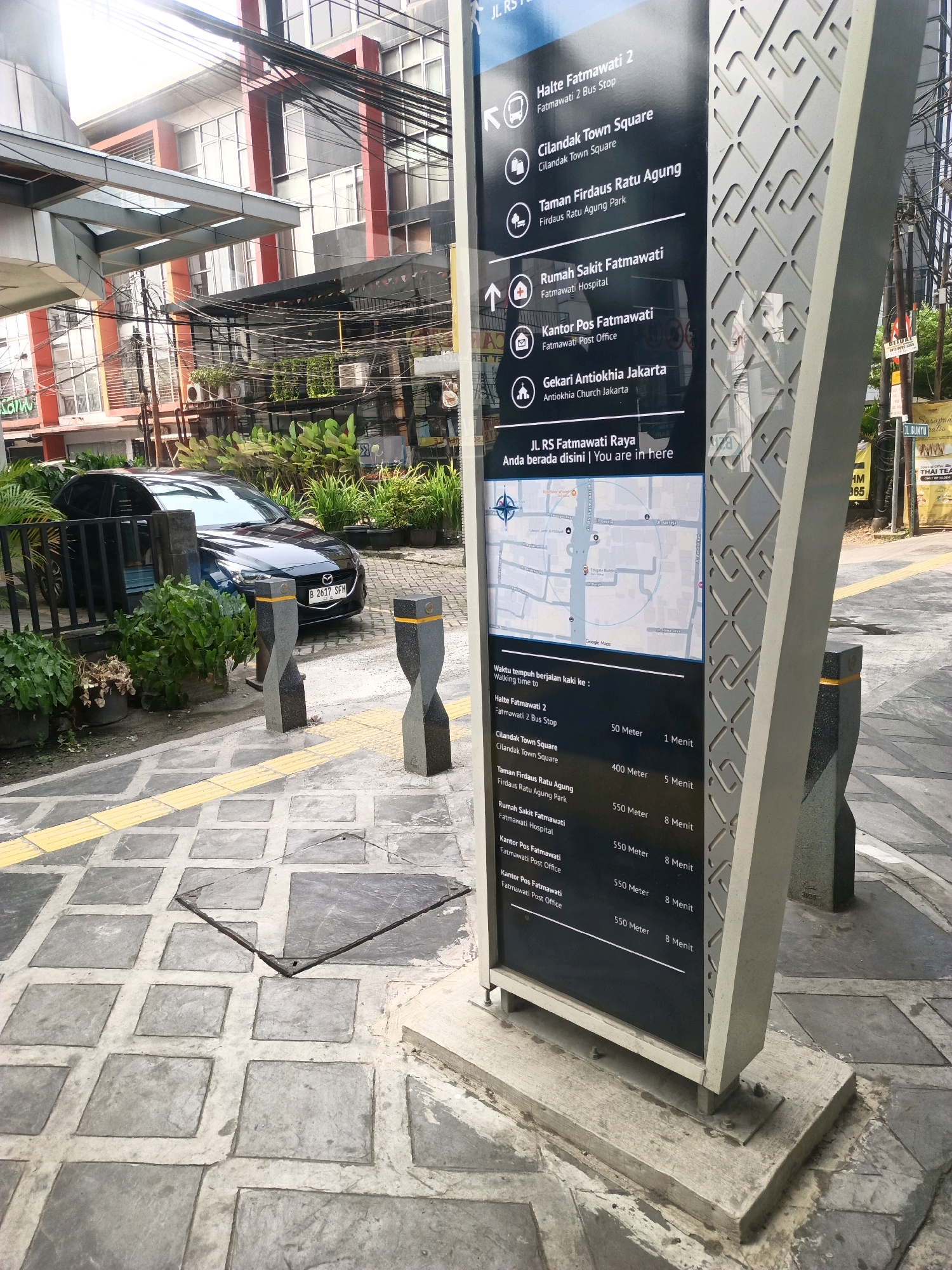 The image shows a directional signpost located on a sidewalk in South Jakarta, Indonesia. The signpost is made of black material with white text and graphics, and it lists several locations with their corresponding distances and estimated walking times. Some of the listed locations include "Halte Fatmawati 2 Bus Stop," "Cilandak Town Square," "Taman Firdaus Ratu Agung," "Rumah Sakit Fatmawati," "Kantor Pos Fatmawati," and "Gekari Antiokhia Jakarta." A map is also displayed on the signpost.
The environment appears to be a street or pedestrian walkway with a paved surface. In the background, there is a building with multiple floors and windows, as well as some greenery and trees. A black car is parked near the sidewalk, and there are concrete bollards on the walkway. The overall scene suggests an urban setting with public transportation and civic amenities. The signpost's prominent placement indicates that it is a point of information for navigating the area.