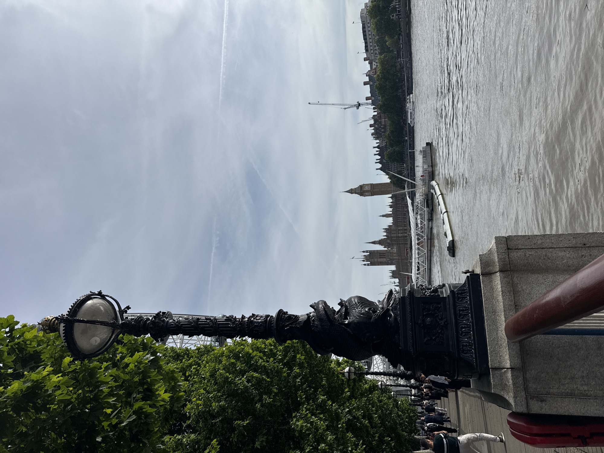 The image depicts a view along the River Thames in London, United Kingdom. In the foreground, a large, ornate black lamppost stands prominently on a granite base. To the left, a tree with dense green foliage lines a paved walkway where several pedestrians are seen walking. The walkway is adjacent to the river, which occupies the right side of the frame. In the background, the Houses of Parliament, including the Elizabeth Tower (Big Ben), are visible across the river. A boat is moving on the water, and a construction crane is visible amongst the buildings. The sky is overcast with some light clouds and contrails from airplanes. The overall scene suggests a typical day on the South Bank of the River Thames, a popular tourist area.