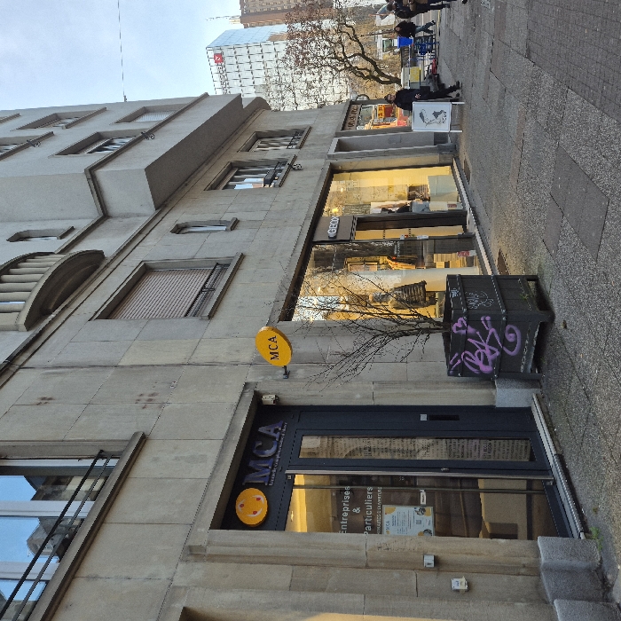 The image captures a street scene from a slightly low angle, looking upwards at the ground floor and lower levels of a building. The perspective is tilted, making the building appear to lean diagonally across the frame.
On the left side, a multi-story building with a facade of light-colored stone or concrete is visible. It features several rectangular windows, some with closed external roller shutters, and others with reflections of the sky or opposite buildings. The architecture includes recessed sections and a decorative, curved, louvered element on one of the upper windows.
The ground floor of this building houses several businesses. Towards the bottom left, a dark-framed glass door is visible, above which is a sign for "MCA". This sign includes a yellow circular logo with a smiley face and the text "MCA" in dark letters. Below "MCA", smaller text reads "Entreprises & Particuliers" and even smaller text underneath is partially obscured but seems to refer to services ("BILANS COMPTOIRS", "FISCALITÉ"). To the right of this door, a circular yellow sign is affixed to the building with the letters "MCA" in black.
Further to the right on the ground floor, a large shop window displays merchandise and has a sign inside that reads "GEOX". The window reflects the street scene opposite, including what appears to be another building and some bare tree branches. In front of this shop window, on the sidewalk, is a dark rectangular planter containing a small, bare tree. The planter has purple graffiti on its side, which appears to spell "Beks" or something similar.
The sidewalk itself is made of light-colored paving stones. Several people are visible walking along the sidewalk towards the right side of the frame, their figures mostly in shadow or indistinct. One person in a dark coat stands near a white A-frame sign on the sidewalk, which features an illustration of two birds.
In the background, behind the building and trees, a modern, glass-clad building with a grid pattern is visible. A large red logo, possibly an "R" or a stylized letter, is prominent on its side against a white background. The sky is bright and appears to be daytime, possibly late morning or early afternoon, given the clear lighting and minimal shadows. The trees are bare, suggesting it is a colder season, like late autumn or winter.