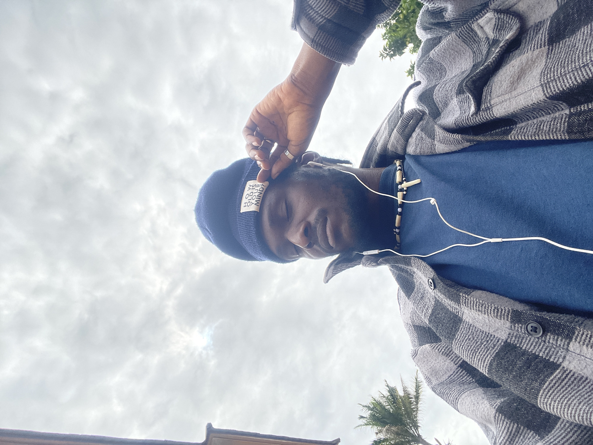 A medium shot captures a Black man from the chest up, wearing a blue beanie with white text that reads "HOW OLD IS YOUR MIND?", a dark blue t-shirt, and a grey and black checkered flannel shirt. He is also wearing a necklace with wooden beads and a cross pendant, and has white earbuds in. The man's eyes are closed, and his right hand is touching his beanie. The background is an overcast sky with clouds, and hints of greenery and a palm tree are visible on the left. The overall lighting is natural and diffused, suggesting an outdoor setting, possibly in Jos, Nigeria, given the presence of the palm tree and the clothing style.