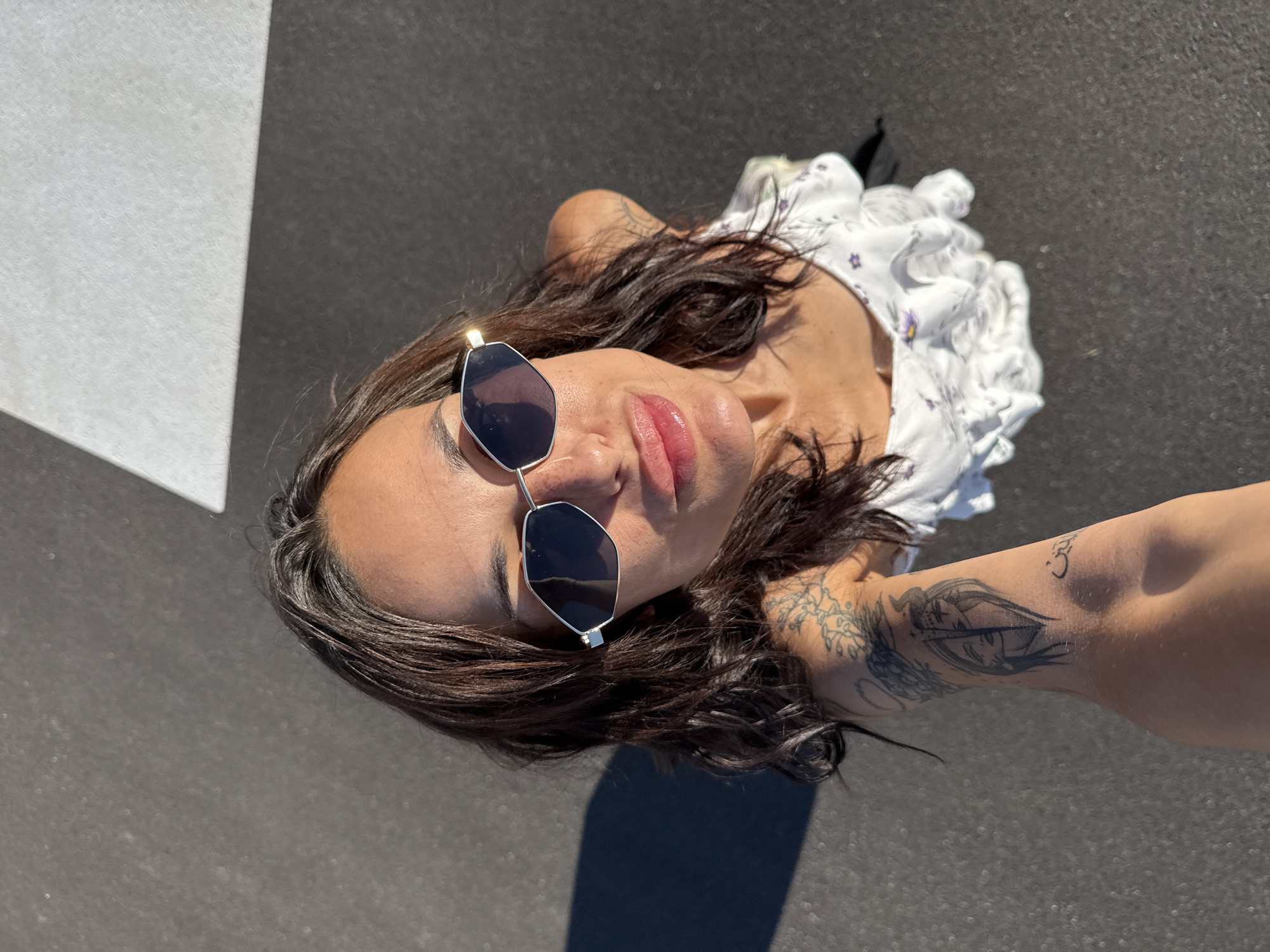 An overhead shot captures a young woman with long, dark, wavy hair, wearing a white floral top and geometric sunglasses. She is looking up at the camera, with her lips slightly parted, and has multiple tattoos on her left arm. The woman is standing on a dark, asphalt-like surface, with a bright white line marking a division in the surface behind her. The lighting suggests it is daytime, with a distinct shadow cast to her left. The scene is set on a street in the city of Pleven, Bulgaria.