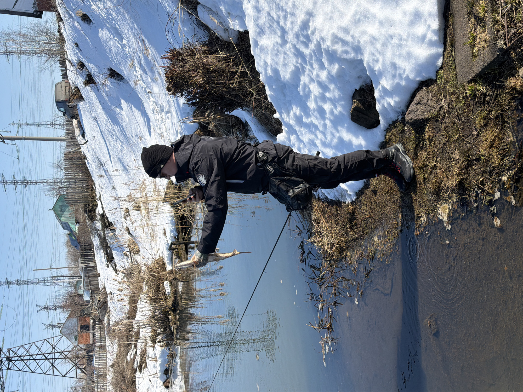 A man in dark clothing and a beanie is lying prone on the bank of a narrow body of water, possibly a small river or stream, with melting snow on the ground nearby. He is holding a stick or branch in his hands, which is partially submerged in the water. A fishing rod is positioned alongside him with its line also entering the water. The water reflects the clear blue sky. On the opposite bank, there are residential houses, power lines, and utility poles, suggesting a location within or near a residential area. The location is likely Chaikovsky, Russia.