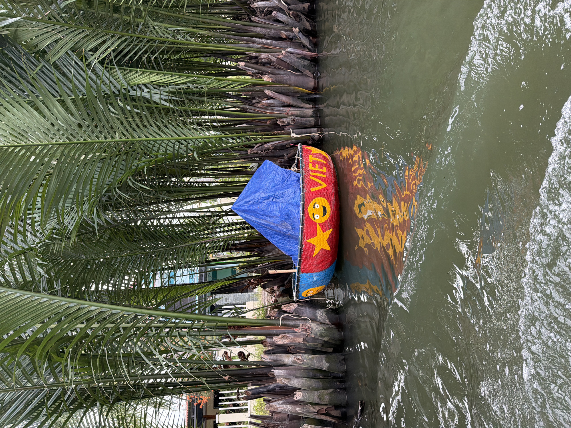 This image shows a small, colorful boat with a blue tarp on a river in Hoi An, Vietnam. The boat is red with yellow and blue accents, and it has a painted smiley face and the word "VIET" on its side. It is surrounded by tall, thin palm trees, and their reflections are visible in the water. The water is green and slightly choppy, with a foamy white edge in the foreground. The scene appears to be a canal or a narrow waterway within the city.