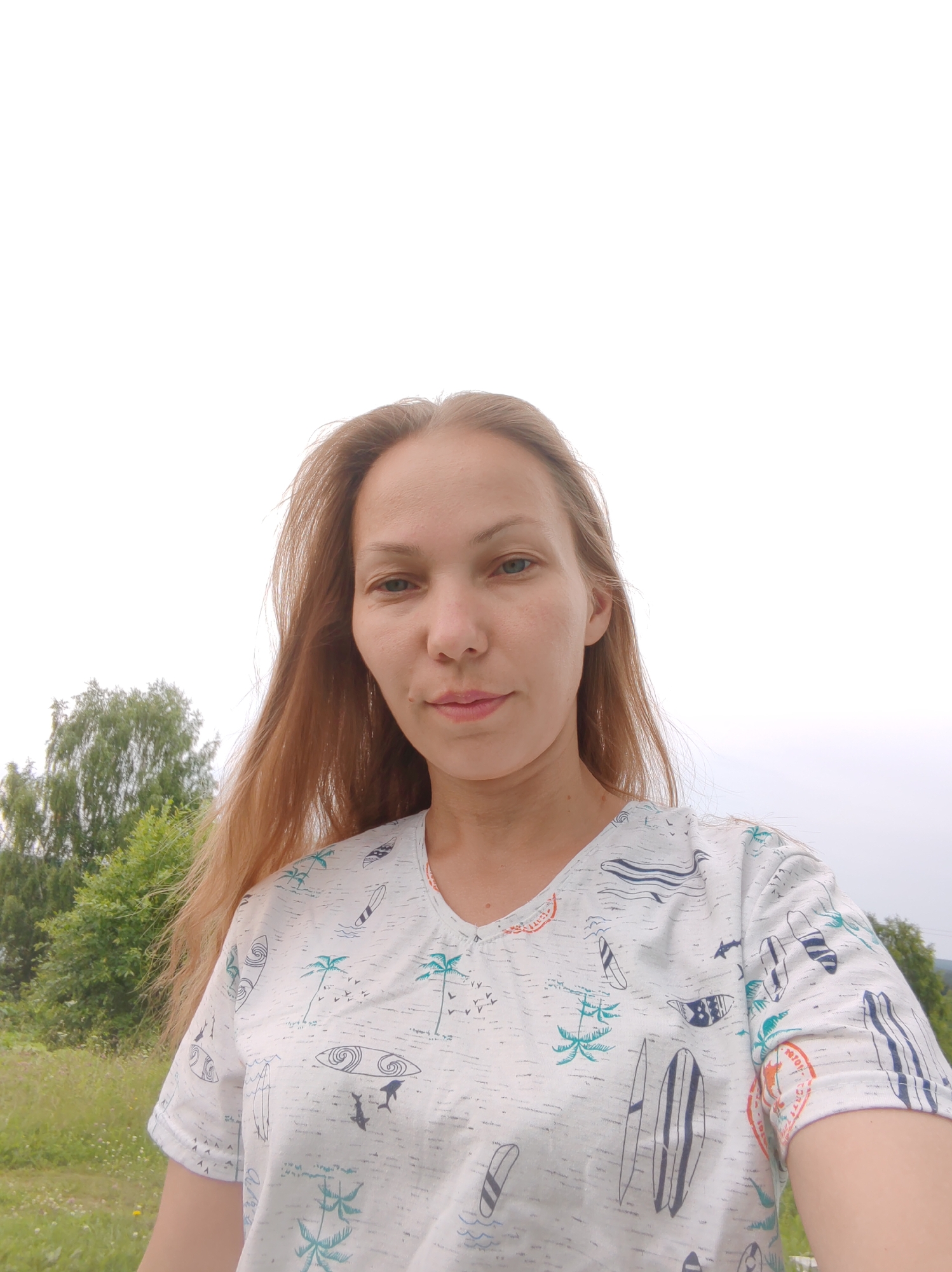 A medium shot taken from a low angle shows a fair-skinned woman with shoulder-length auburn hair standing outdoors. She is wearing a white t-shirt with a graphic print of surfboards, palm trees, and seagulls. The woman is looking towards the camera with a neutral expression. The background consists of green trees and grass, under an overcast sky. The image appears to have been taken in a rural or park-like setting. There is no specific indication of the location within the city of Чухлэм, Russia, other than that the natural environment suggests a possible outdoor area within or near the city.