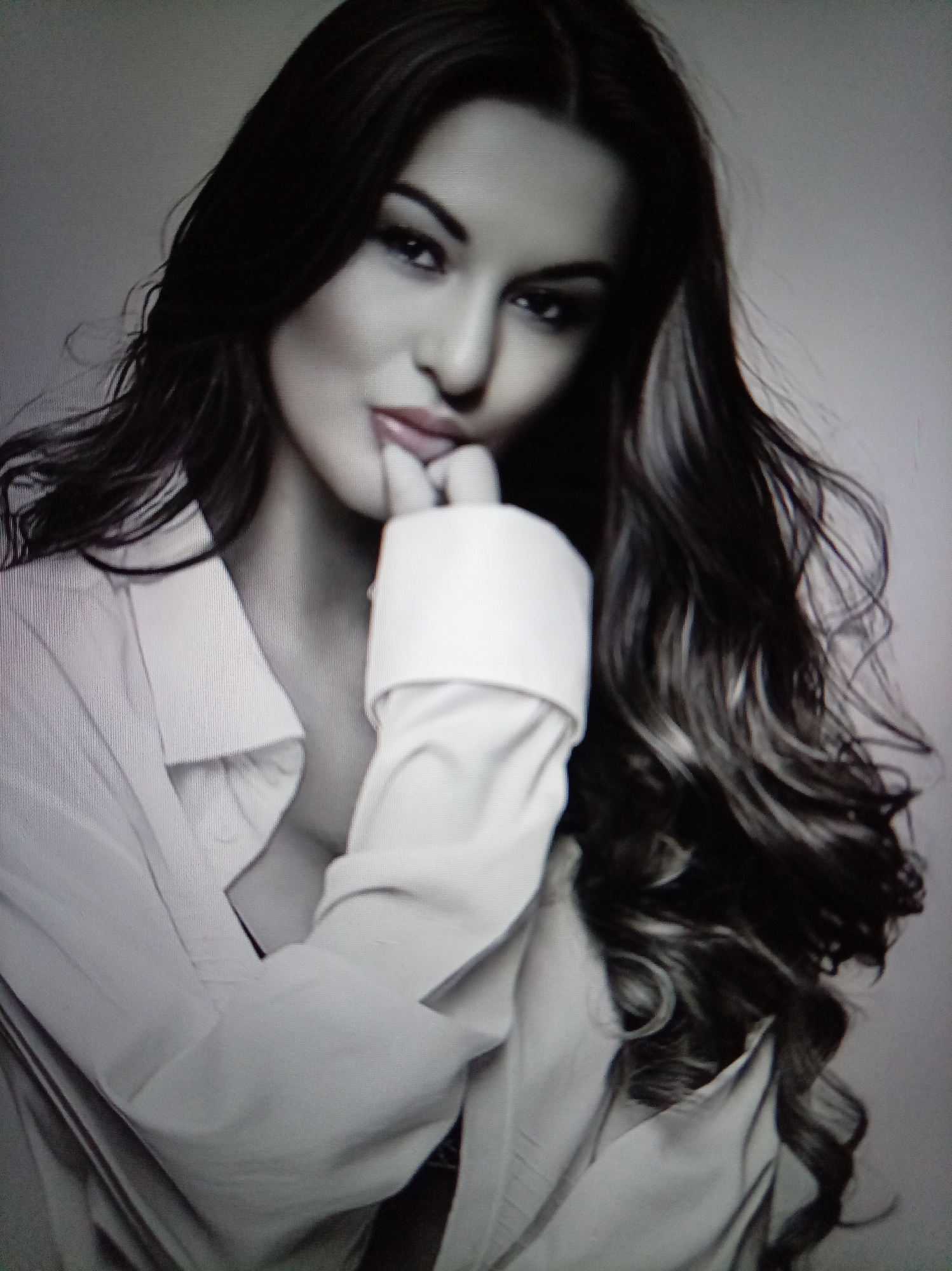 A close-up, black and white studio portrait shows a woman with dark, wavy hair and fair skin. She is wearing a white button-down shirt, unbuttoned, revealing a glimpse of dark clothing underneath. Her left hand is raised to her lips, with her index finger gently touching her lower lip. Her gaze is directed towards the viewer. The background is a plain, light gray wall. There is no indication of the city of Yessentuki, Russia, in the image.