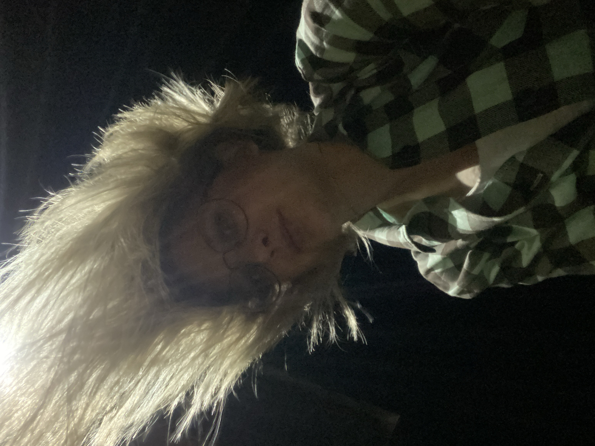 A person wearing round-rimmed glasses, a fur-trimmed hooded jacket and a plaid shirt stands outdoors at night. The location is unknown.