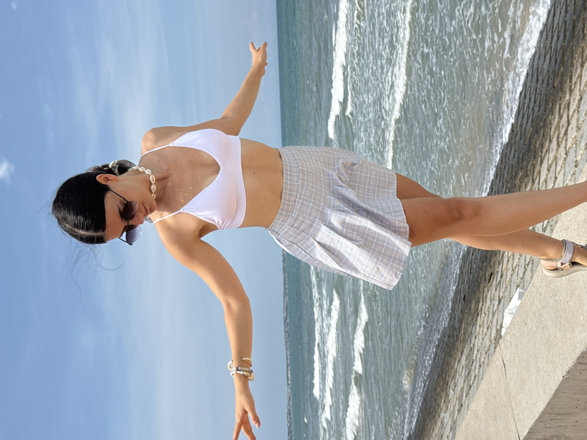 A young woman with dark hair styled in a braid and wearing a white bikini top, a pale blue plaid skirt, sunglasses, and a seashell necklace stands on a concrete ledge overlooking the sea in Phu Thuy Ward, Vietnam. Her arms are outstretched, and she is looking down. The ocean is blue with white-capped waves, and the sky is clear and blue with a few wispy clouds. The concrete ledge is rough and textured, and there is a rough stone embankment behind it.