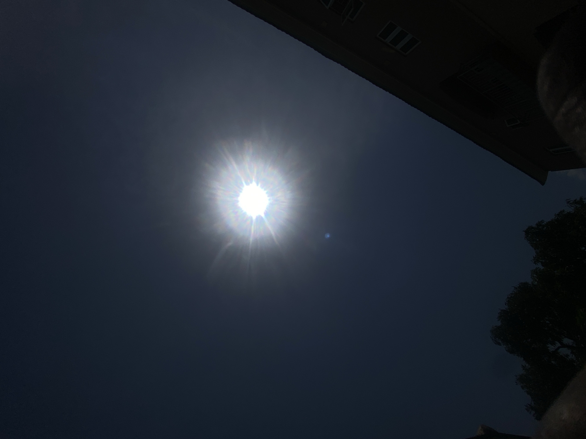 The image shows the sun in the sky, seemingly partially obscured by cloud cover or atmospheric haze, with visible solar flares emanating from its center. A portion of a building or structure is visible in the upper right corner of the frame, and what appears to be the top of a tree is visible in the lower right. The location and city details requested are not discernable from the image. No people, subjects, objects, actions, or interactions are visible, save for the celestial ones.